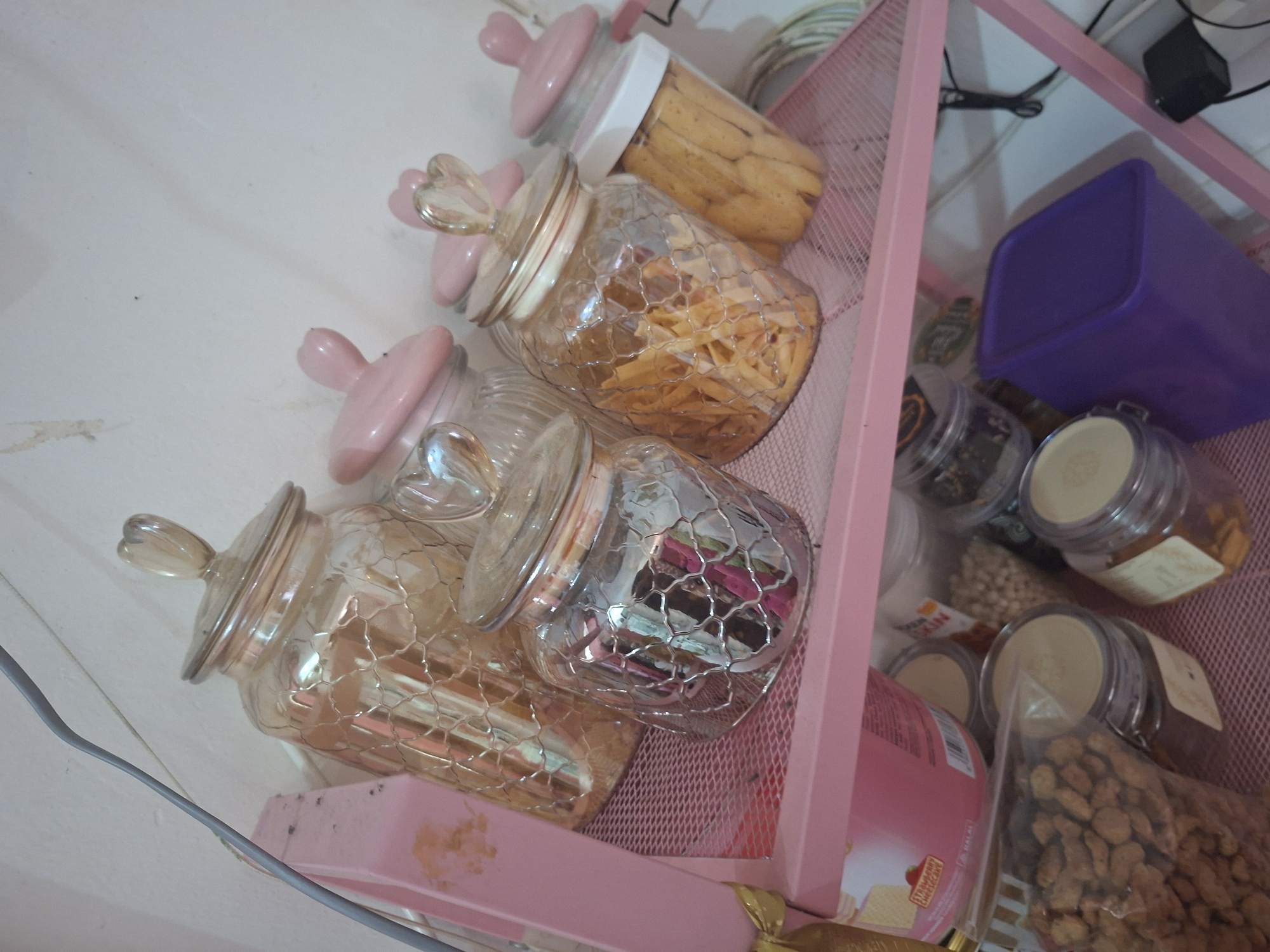 The image captures a collection of food storage containers, primarily glass jars, arranged on pink mesh shelves in what appears to be an indoor setting, likely a kitchen or pantry.
In the foreground and central part of the image, several decorative glass jars are visible. These jars feature distinctive pink lids, some adorned with heart-shaped glass handles. The glass bodies of these jars have an intricate, possibly chicken wire-like or cracked, textured pattern. They contain various snacks: one jar holds light-brown, elongated biscuits, while another contains irregularly shaped, fried golden snacks.
On the pink mesh shelves, other storage items are arranged. A solid purple rectangular container with a lid sits on a lower shelf. Several smaller, round glass jars with light-colored or beige lids are also present, containing various contents such as small, round, white candies or nuts, and what appears to be other types of snacks or spices. A clear plastic bag filled with peanuts or similar small, brown snacks is also visible on the bottom right.
A pink box or package with "STRAWBERRY" and "HALAL" printed on it is partially seen on the lower shelf. Another label on a smaller jar partially shows "TOP GULINDO". A pink item with "SKIN" is also faintly visible on another package. Cables or wires are visible in the upper background, contrasting against a white wall. The overall scene suggests a tidy, organized storage area with a pastel pink aesthetic, common in homes in Ciseeng, Indonesia.