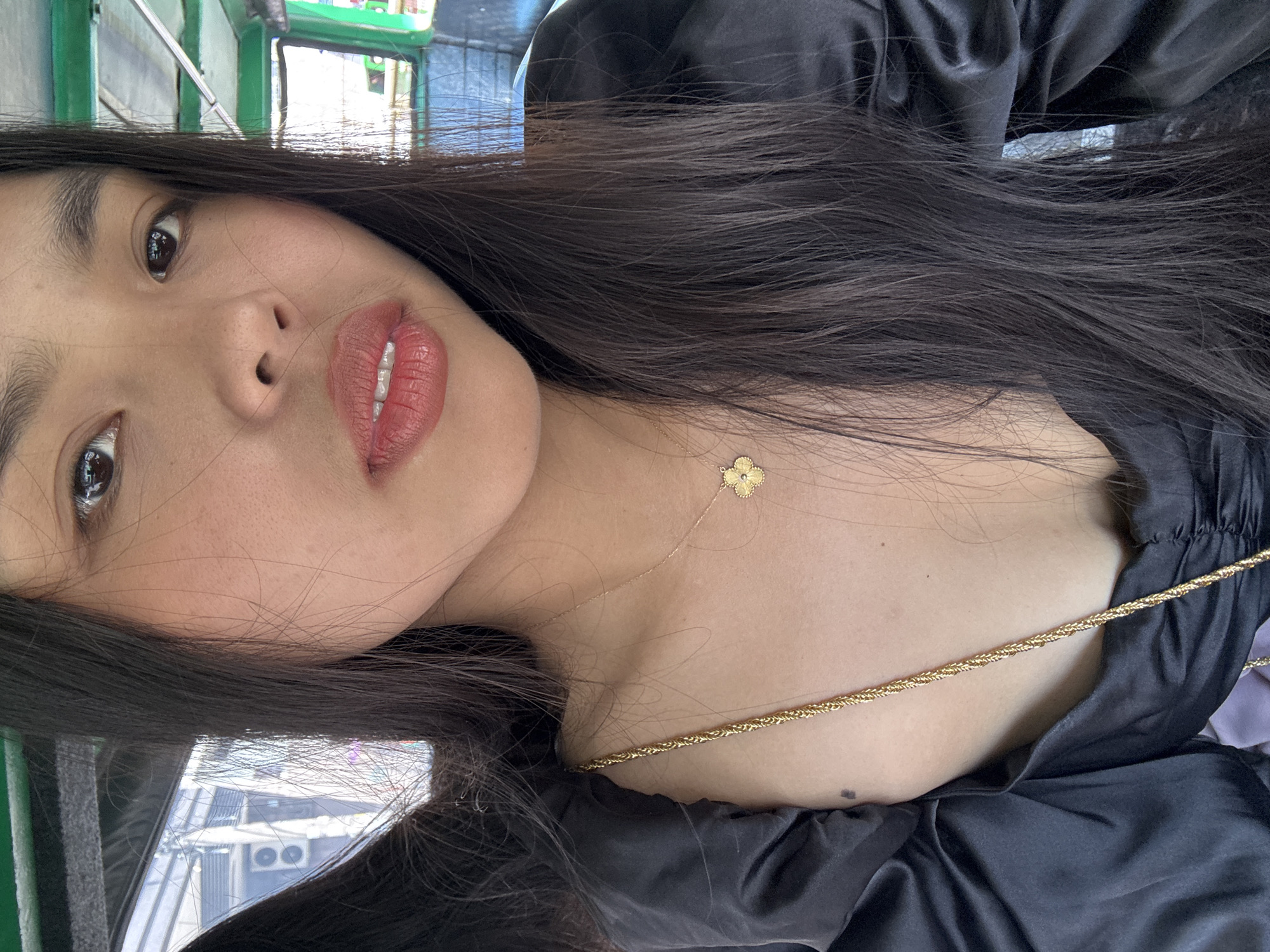 The image is a close-up portrait, rotated approximately 90 degrees clockwise, featuring a young woman looking directly at the camera. Her face, positioned centrally, shows clear skin, dark eyes, and full lips enhanced with a matte reddish-brown or terracotta-colored lipstick. Long, dark, straight hair cascades over her shoulder and chest.
She is wearing a black top or dress with a satin-like texture, which creates soft folds and appears to have a wide or off-shoulder neckline. Adorning her décolletage is a delicate gold-colored chain necklace featuring a small, four-leaf clover or flower pendant. A thicker, braided gold-colored chain, possibly a bag strap or another piece of jewelry, diagonally crosses her upper chest.
The background is largely out of focus, providing subtle cues of an outdoor or semi-enclosed setting. A green structure or railing is partially visible in the upper left, and reflections in her eyes and through her hair suggest urban elements like buildings or air conditioning units. The lighting indicates it is daytime. The woman maintains a calm and direct gaze. No visible text is present in the image, which was taken in Davao City, Philippines.