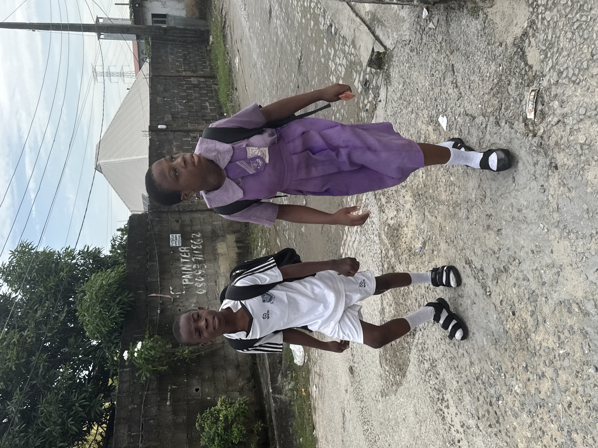 Two children stand on a rough, paved street in what appears to be an urban or semi-urban setting in Ado, Nigeria. The scene is brightly lit by natural daylight, suggesting it is daytime, under an overcast sky.
The child on the left is a girl, wearing a light purple short-sleeved school uniform shirt with a collar, a darker purple pleated skirt, white socks, and black and white sandals. She carries a black backpack. A logo or crest is visible on the pocket of her shirt. She looks towards the camera with a neutral expression.
The child on the right is a boy, dressed in a white and black short-sleeved sports uniform, including a white t-shirt with black accents on the sleeves and collar, and white shorts. He also wears white socks and black and white sandals, which feature the Adidas logo on the straps. He carries a black backpack and looks towards the camera with a slight smile. The text "G SPORTS" is visible on his shirt and shorts.
The background features a low concrete block wall partially covered by lush green trees and foliage on the left. Further back, buildings with corrugated roofs are visible, along with utility poles and power lines crisscrossing the sky. A large electricity pylon stands in the distance. On a wall behind the boy, white painted text reads "PAINTER" followed by phone numbers "08053 711862" and another partially obscured number beginning with "070630". The ground is a mix of concrete and gravel, with some debris scattered about.