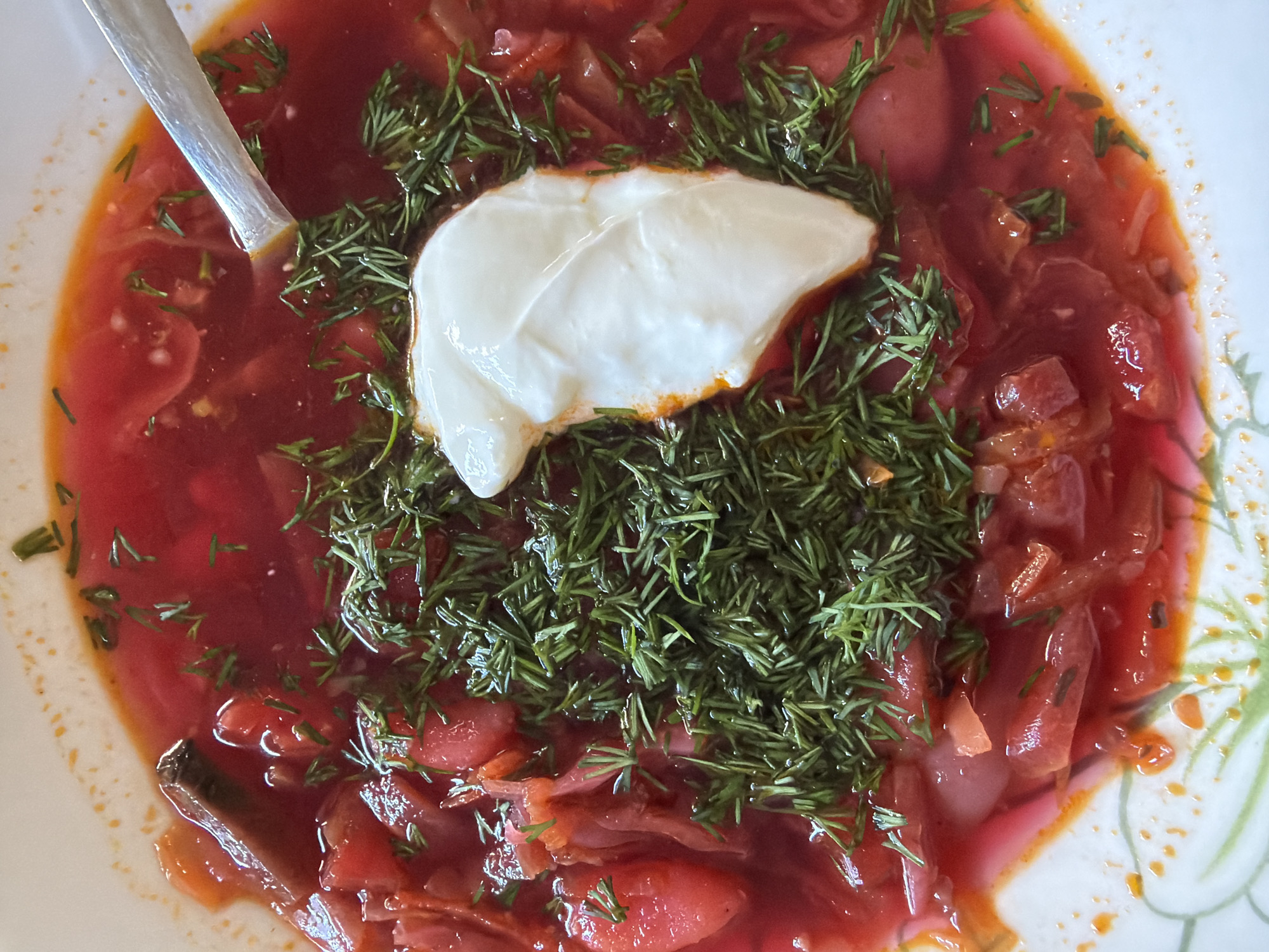A close-up shot captures a bowl of borscht, a vibrant red soup, topped with a dollop of white sour cream and fresh dill. A silver spoon rests in the upper left corner of the bowl. The borscht contains visible pieces of beets and other vegetables. The bowl is white with a faint floral pattern along the edge. The location is unknown, and there are no people present in the image.