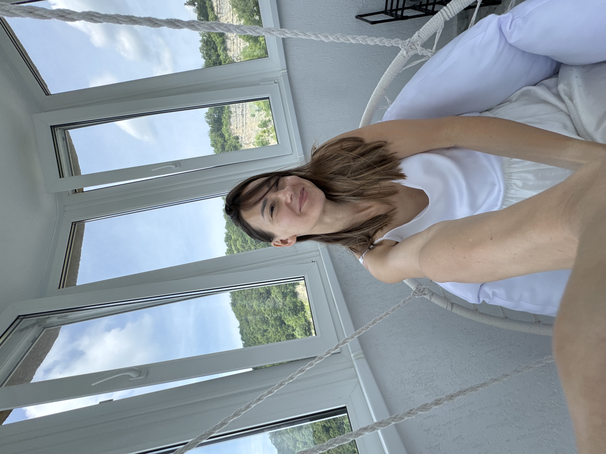 A woman is seated in a white hanging chair on a balcony in Sochi, Russia. She is wearing a white tank top and white skirt. The balcony has large windows that overlook a lush green hillside with a rocky cliff face. The sky is cloudy. She is holding a phone in her right hand, taking a selfie. The chair is suspended by thick white ropes.