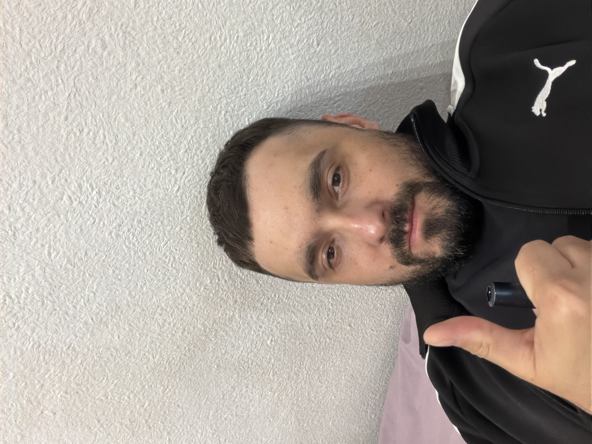 A man with short, dark hair and a beard is posing for a selfie indoors. He is wearing a black Puma jacket with white accents. He is holding a small black object in his hand and giving a thumbs-up. The background is a textured white wall. The location in the city of Kovel, Ukraine is not discernable from the image.