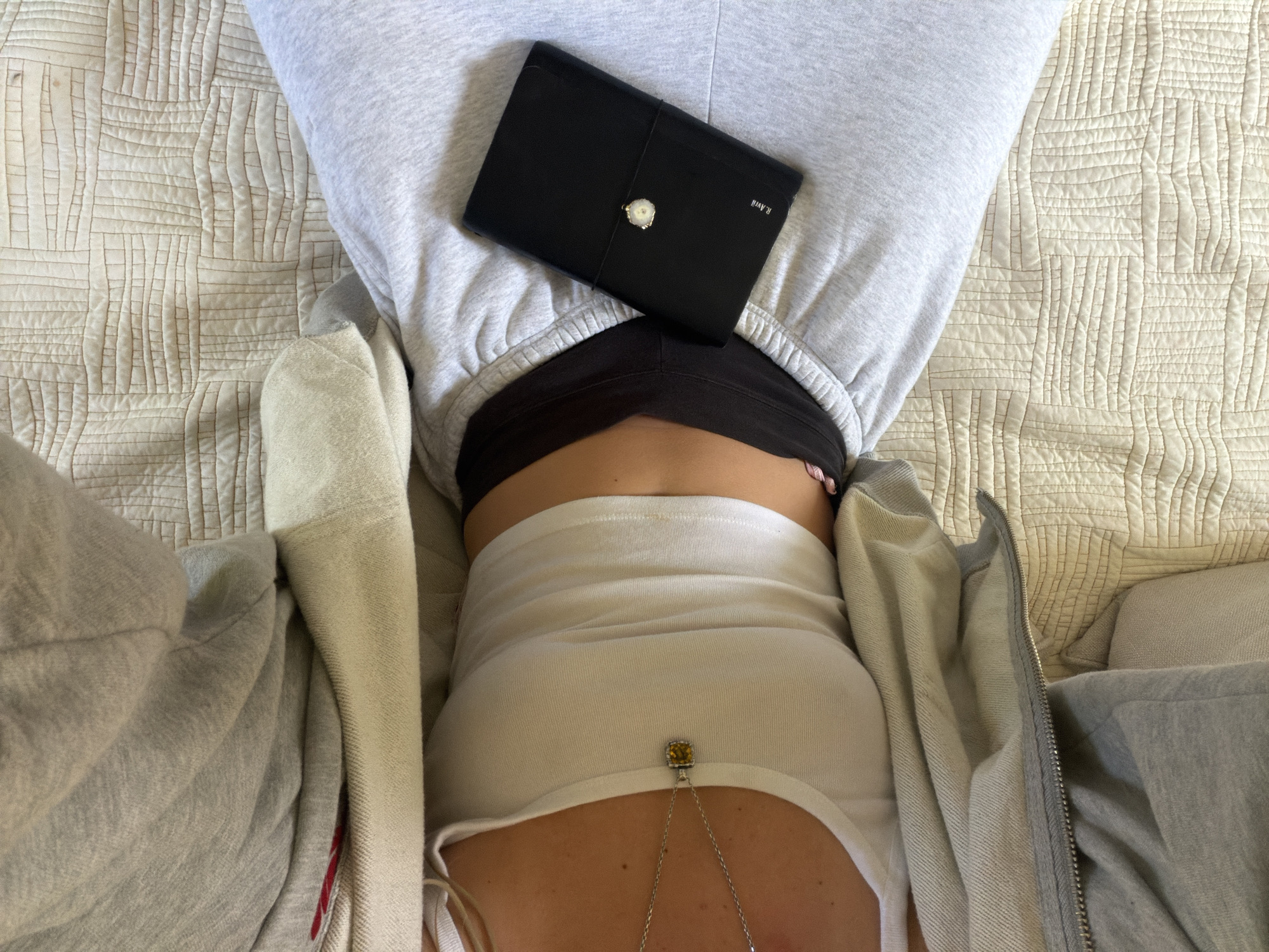 An overhead shot captures a person lying on their back, likely on a bed or sofa, within an indoor setting in Los Angeles, United States of America. The image focuses on the person's torso and lower body, dressed casually in a light gray hooded sweatshirt, a white tank top underneath, and a black sports bra or top visible beneath the sweatshirt's hood.
A black planner or notebook rests on the person's upper abdomen, specifically on the gray sweatshirt. This item features a distinctive white and silver decorative clasp, possibly a geode or crystal, positioned centrally on its front. The text "R. Avrii" is visible in white lettering on the right side of the planner.
A silver chain necklace with a prominent square-shaped yellow or amber-colored pendant, possibly a gemstone, hangs from the person's neck, extending down towards their navel area and resting on the white tank top.
The person is situated on a light cream or beige quilted bedspread or blanket, which displays a geometric pattern of raised squares and vertical lines across its surface. The scene conveys a sense of comfort and relaxation in a personal space.