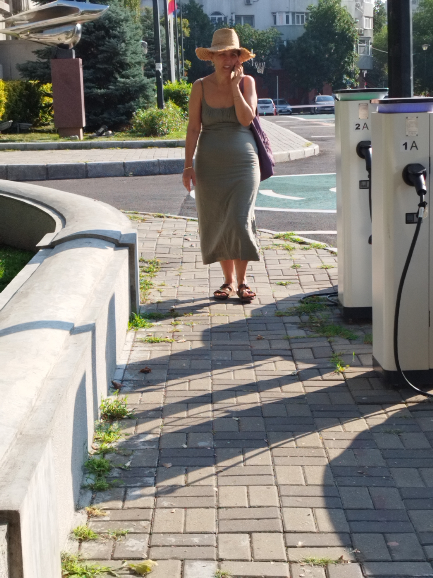 A woman wearing a straw hat and a long, olive-green dress is walking on a paved sidewalk in Bucharest, Romania. She is holding her phone to her ear, as if in a conversation. She has a purple bag slung over her shoulder. The sidewalk is made of grey rectangular pavers and is lined with patches of grass and weeds. In the background, there are several electric vehicle charging stations, labeled "2A" and "1A." Behind the charging stations, there is a street with cars, and beyond that, buildings and trees. To the left of the woman, there is a curved concrete structure. The lighting suggests it is daytime, with strong shadows cast by the woman and the surrounding objects.
