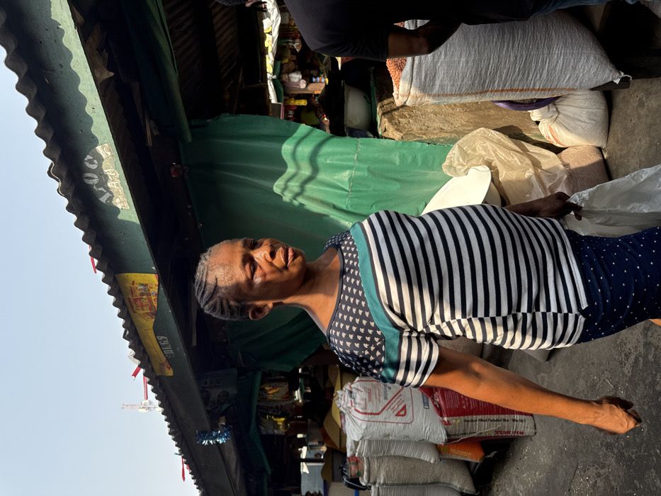 The image displays an outdoor market scene in Lekki, Nigeria, under bright daylight. A woman of African descent is centrally positioned, walking towards the right of the frame with a neutral expression. She has braided hair and wears a short-sleeved top with a dark blue patterned upper section, a teal horizontal stripe, and a main body of black and white horizontal stripes. Her lower garment is dark blue with light polka dots. She carries a white plastic bag in her right hand. To her right, the back and shoulder of another individual, wearing a dark short-sleeved shirt, are partially visible.
In the background, market stalls with green corrugated metal roofs are visible, adorned with red plastic strips and blue tinsel. Green tarpaulin or fabric serves as backdrops for the stalls. Numerous large white and off-white sacks, likely containing dry goods, are stacked on both sides of the scene. Smaller bags are also present on the ground. A construction crane is faintly visible in the far upper background against a clear sky. The ground appears to be paved or concrete.
