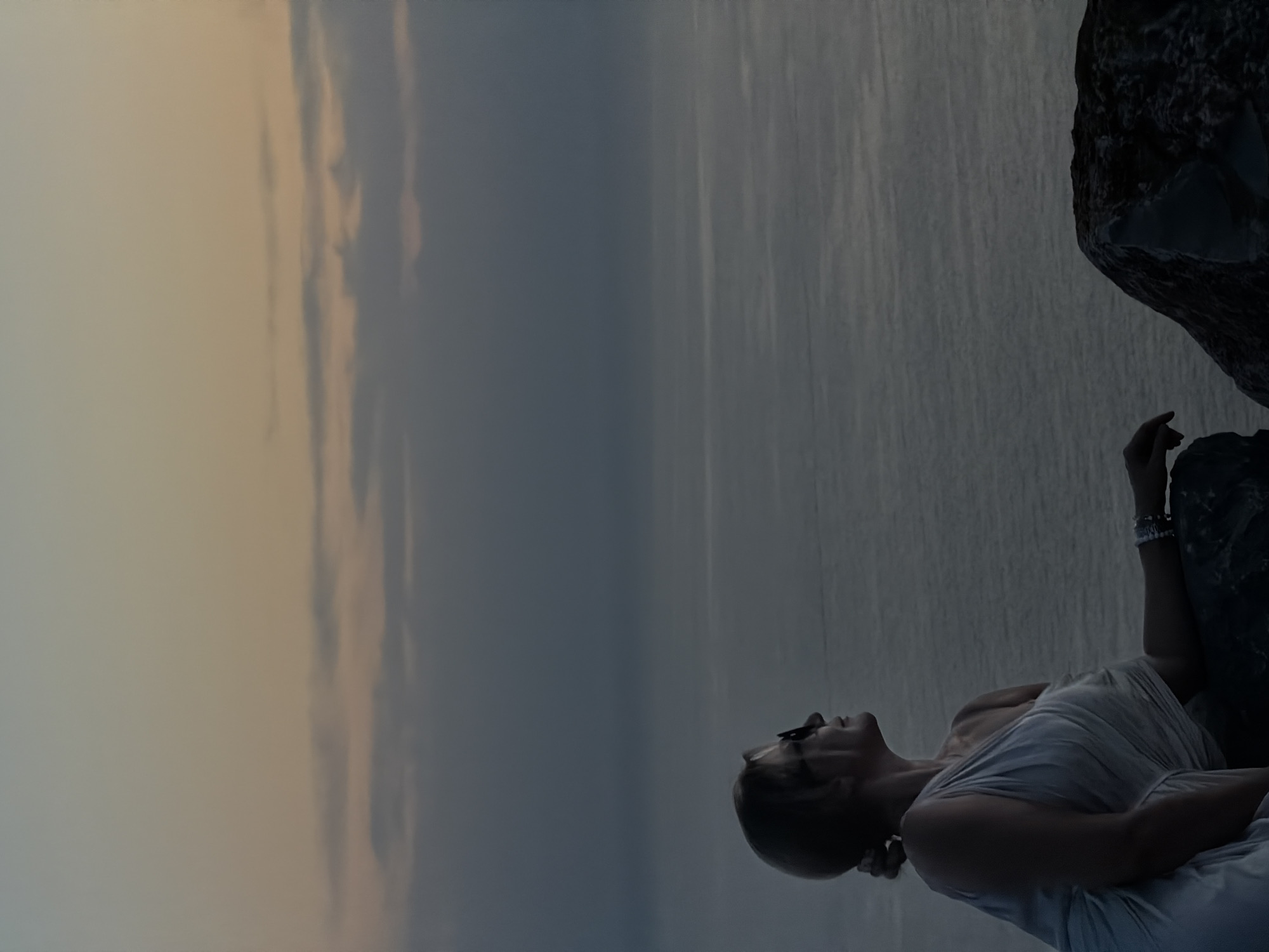 A woman with blonde hair pulled back in a bun and wearing sunglasses is seated on a rocky outcrop, looking out at the sea. She is wearing a light-colored, one-shoulder dress. Her right arm is resting on her leg, and her left arm is extended slightly. The sea is calm, and the sky above is a gradient of pale orange and gray, indicating it is either dawn or dusk. The location is identified as Kovačko Polje, Montenegro.