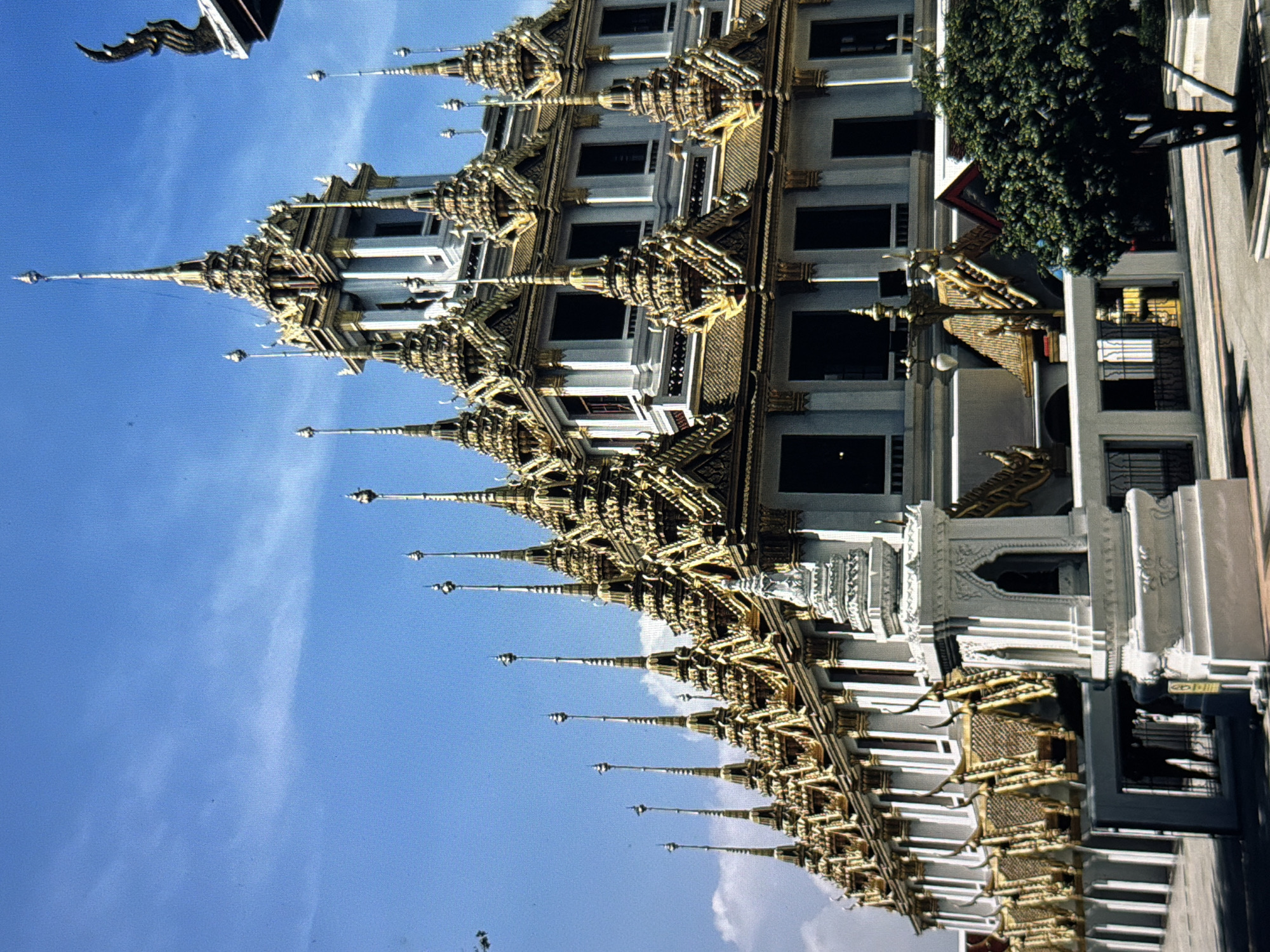 The image shows a tall, ornate building with intricate golden decorations and spires against a blue sky. The building has white walls and numerous windows, some of which are dark. The architectural style appears to be Southeast Asian, possibly Thai. A tree is visible on the right side of the image. The photo is taken from a low angle, looking up at the building. The location of the image is not in Đà Nẵng, Vietnam. It is most likely in Bangkok, Thailand.