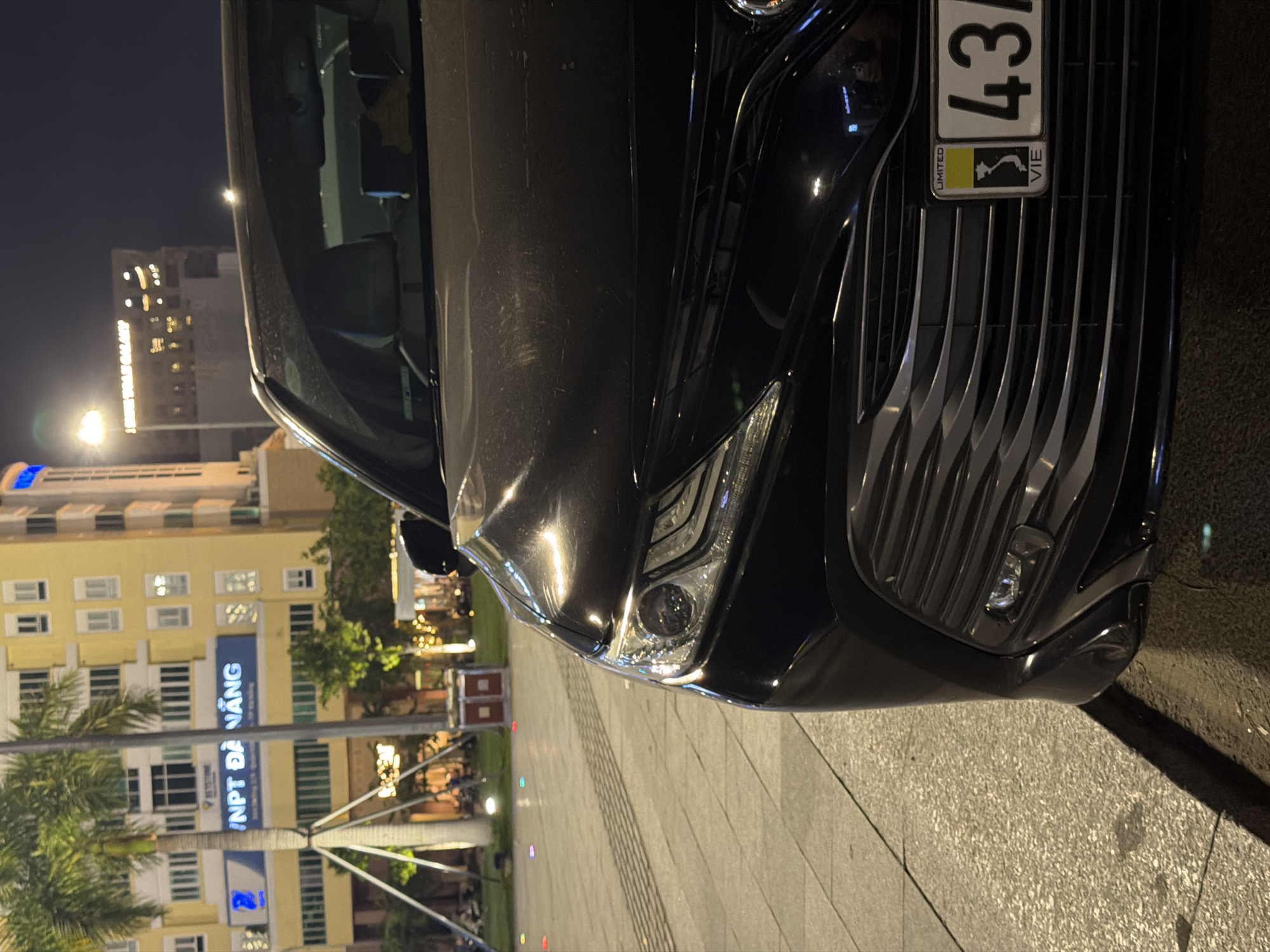 The image shows the front of a black sedan parked on a paved surface in the city of Đà Nẵng, Vietnam. The car is positioned at an angle, with its right headlight and grille prominently visible. In the background, a modern yellow building with multiple windows is illuminated, featuring a blue sign that reads "INPT ĐÀ NẴNG". Palm trees and streetlights are also visible, suggesting an urban nighttime setting. The car's license plate is partially visible, displaying the numbers "43" and the text "VIE". The ground is a light-colored paver stone with a textured pattern.