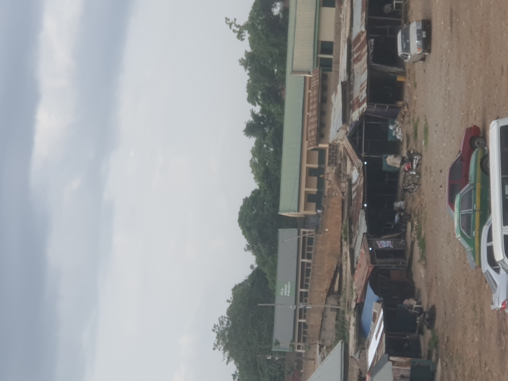 The image depicts a street scene in Jos, Nigeria, characterized by a mix of permanent structures and makeshift stalls. In the background, a long, light-colored building with a green roof is visible, possibly a school or administrative building, with the text "PTA MIDDLE PRIMARY" discernable on its facade. In front of this building and to its right are lower-profile structures, some with corrugated tin roofs, suggesting a marketplace or commercial area.
Closer to the foreground, a row of shops with various roofing materials, including corrugated metal and tarpaulin, are arranged along a dirt ground. Several vehicles are present: a silver Mercedes-Benz sedan parked in front of one of the shops, a green and white sedan further back, and the front of a white van. Two motorcycles are also visible, one with a person in a beige jacket and red pants standing next to it, appearing to be about to mount or dismount. Another person is seated in a white chair outside one of the stalls. The environment is dusty and somewhat unkempt, typical of many market areas in the region. The sky is overcast with gray clouds.