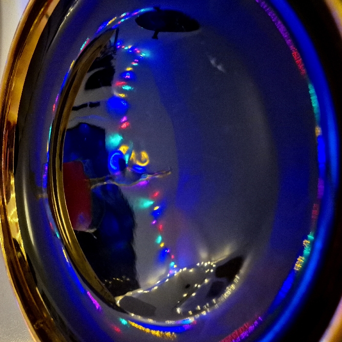 A glossy, gold-rimmed plate reflects a chaotic scene of colorful Christmas lights. The lights are scattered, creating vibrant dots and streaks within the plate's surface. A dark umbrella-like object hangs above, its reflection distorted. The overall effect is one of shimmering, fragmented light captured within the circular frame of the plate. The scene's complexity is enhanced by multiple layers of reflection and the play of light across the plate's reflective surface.