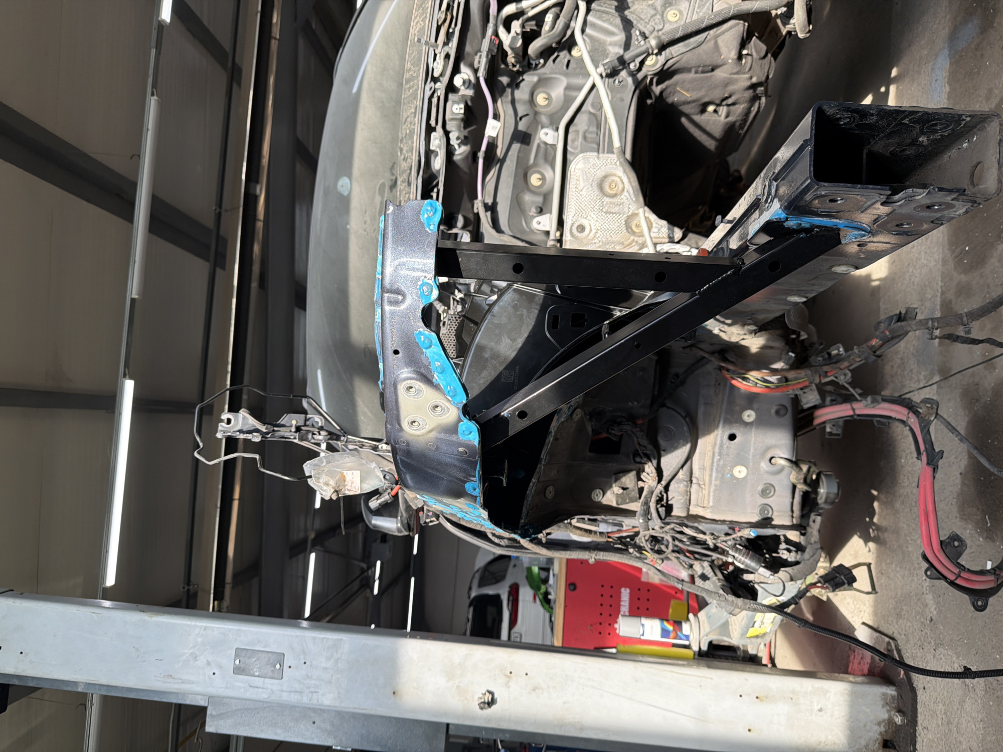 The image depicts the front end of a car undergoing repairs in what appears to be an auto body shop. The vehicle is elevated on a lift. The front body panels have been removed, exposing the engine compartment, wiring, and supporting structures. A black support structure is attached to the front of the car. Tools and equipment are visible in the background, including a red toolbox. The location is potentially a repair shop in Burgas, Bulgaria, but this is not definitively proven from the image itself.