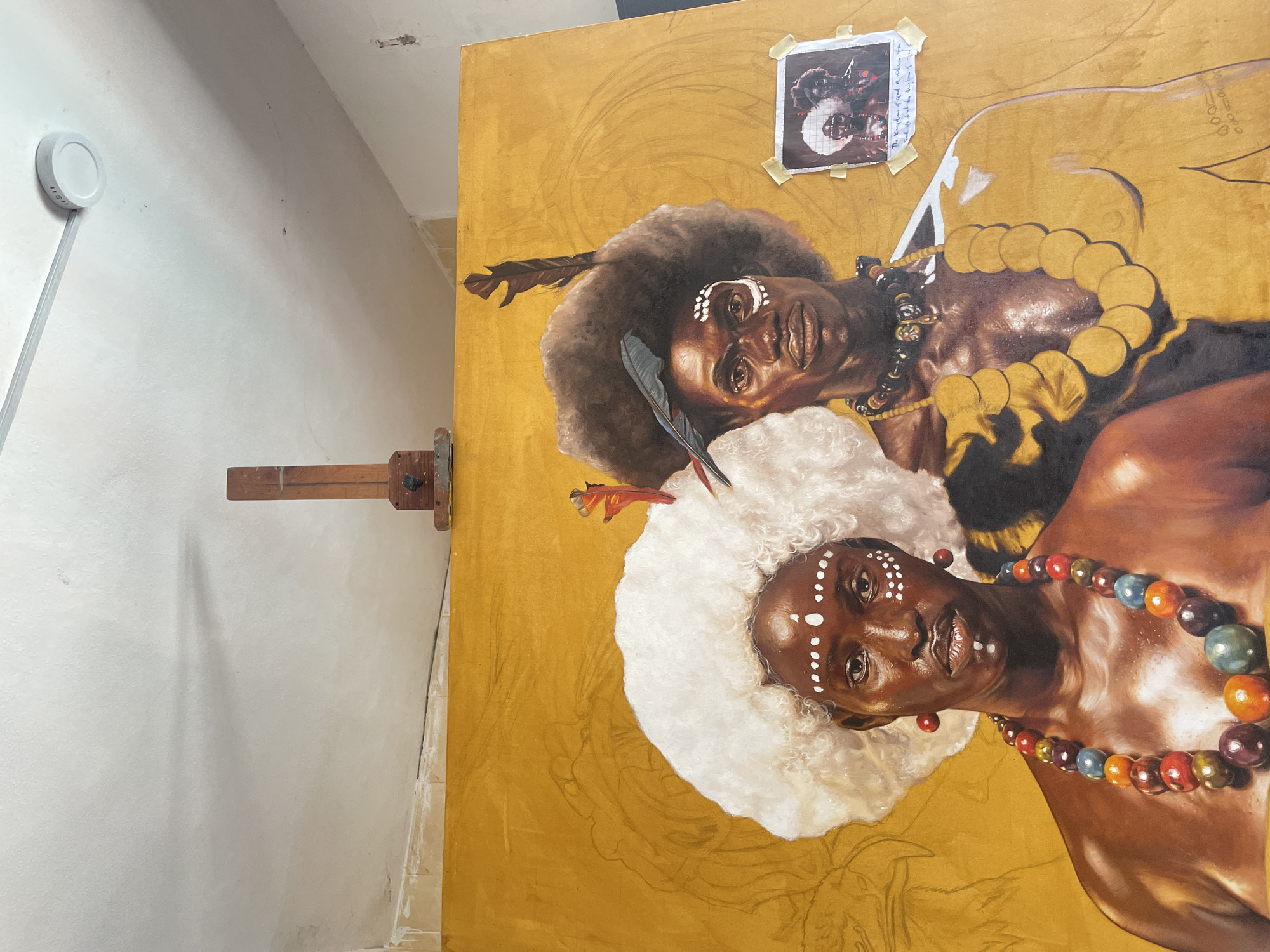 The image shows a portion of an oil painting in progress. The canvas is set up on a wooden easel in a room with light-colored walls. Two figures, depicted from the shoulders up, dominate the painting. Both figures appear to be Black. The figure at the bottom has a white, textured headpiece or collar, white dots painted on the forehead and cheeks, and wears a colorful beaded necklace. The figure at the top has an Afro with feathers and black-and-yellow beaded necklaces. A photograph of the two figures has been taped to the upper right corner of the painting as a reference. There are penciled outlines of a floral design on the gold background of the painting. A white smoke detector is seen on the wall to the left. No indication of the location in Ado, Nigeria is present in the image.