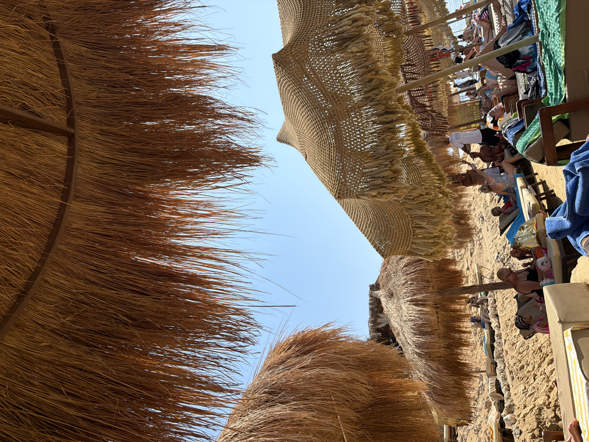 The image depicts a sun-drenched beach in Sharm El Sheikh, Egypt, likely from a resort. A low-angle perspective looking upwards reveals several thatched and woven umbrellas providing shade over a sandy and rocky beach area. People are visible relaxing on loungers, some sunbathing, others conversing. The sky is clear and blue. The umbrellas are made of natural materials, with dried palm leaves for the thatched ones and woven fibers for the others, both featuring decorative fringes. In the background, the sea is visible, suggesting proximity to the water. The overall scene conveys a typical beach resort atmosphere, characterized by leisure and relaxation under the Egyptian sun.