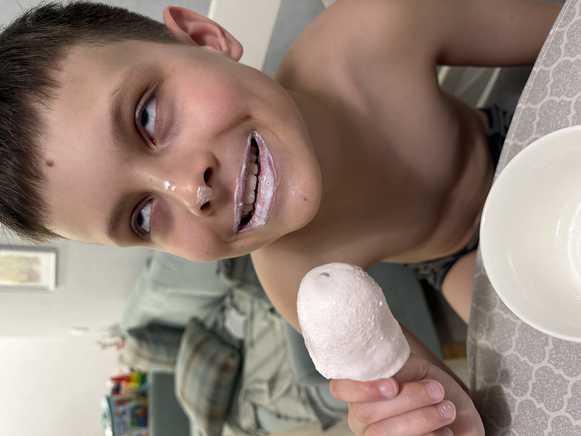 A boy with short brown hair and light skin is holding a white, dome-shaped item, possibly food, in his hand. He has white substance on his lips and nose. He is shirtless, and the lower part of his body is partially visible, revealing patterned underwear. A white bowl sits on a gray patterned tablecloth. In the background, there is a gray sofa with patterned pillows, and a shelf with colorful items is visible. The location in Votkinsk, Russia, cannot be determined from the image.