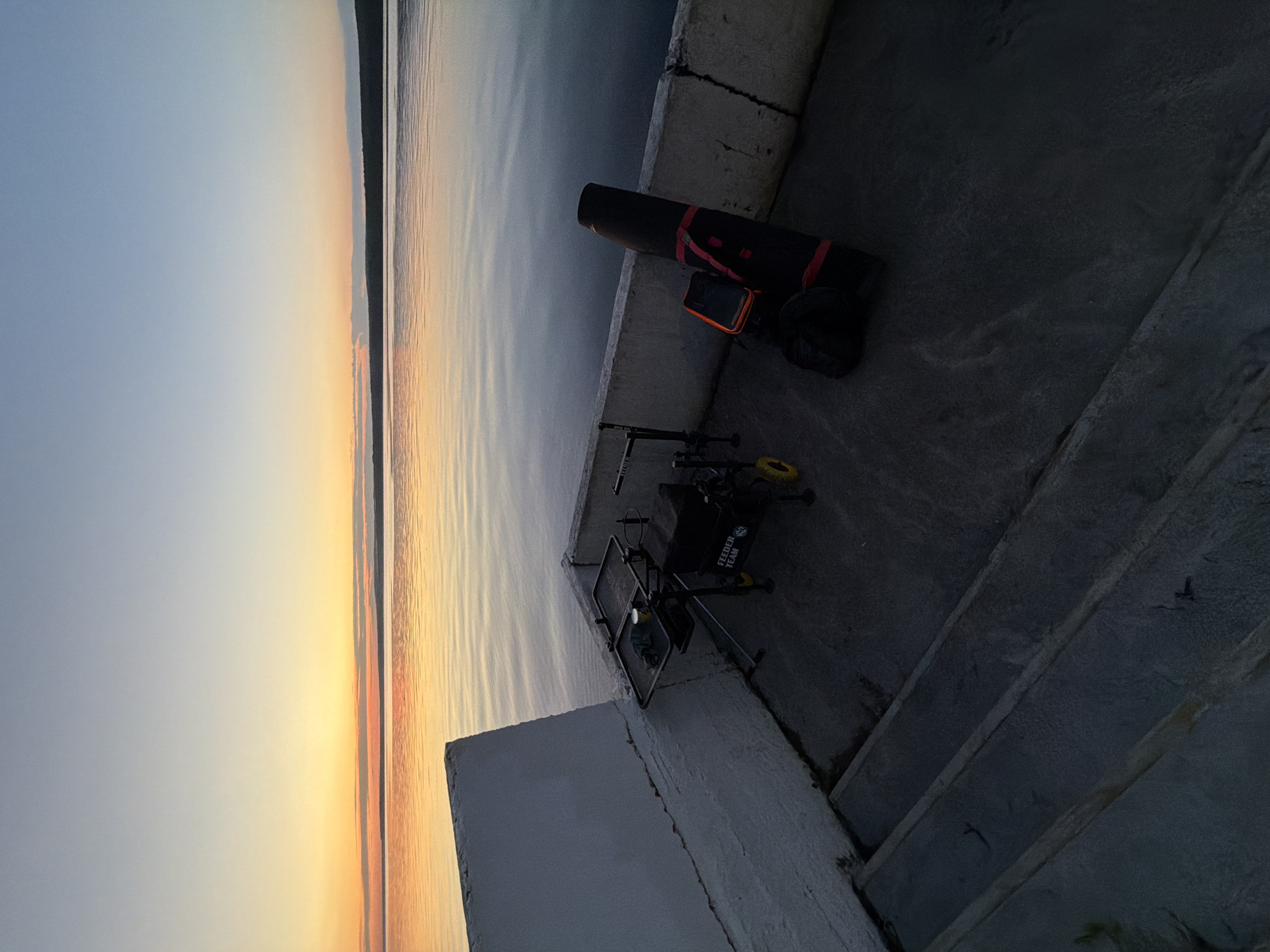 The image depicts a serene sunset scene over a calm body of water, likely the Kama River in Chaikovsky, Russia. In the foreground, a concrete platform or embankment is visible, leading down a set of concrete steps. On the platform, fishing gear is set up, including a feeder rod support system with a black box and yellow wheels, and a large black fishing rod bag with red accents. The sky transitions from a light blue at the top to warm hues of orange and yellow near the horizon, indicating the sun is setting or has just set. The water reflects the colors of the sky, creating a peaceful and tranquil atmosphere.