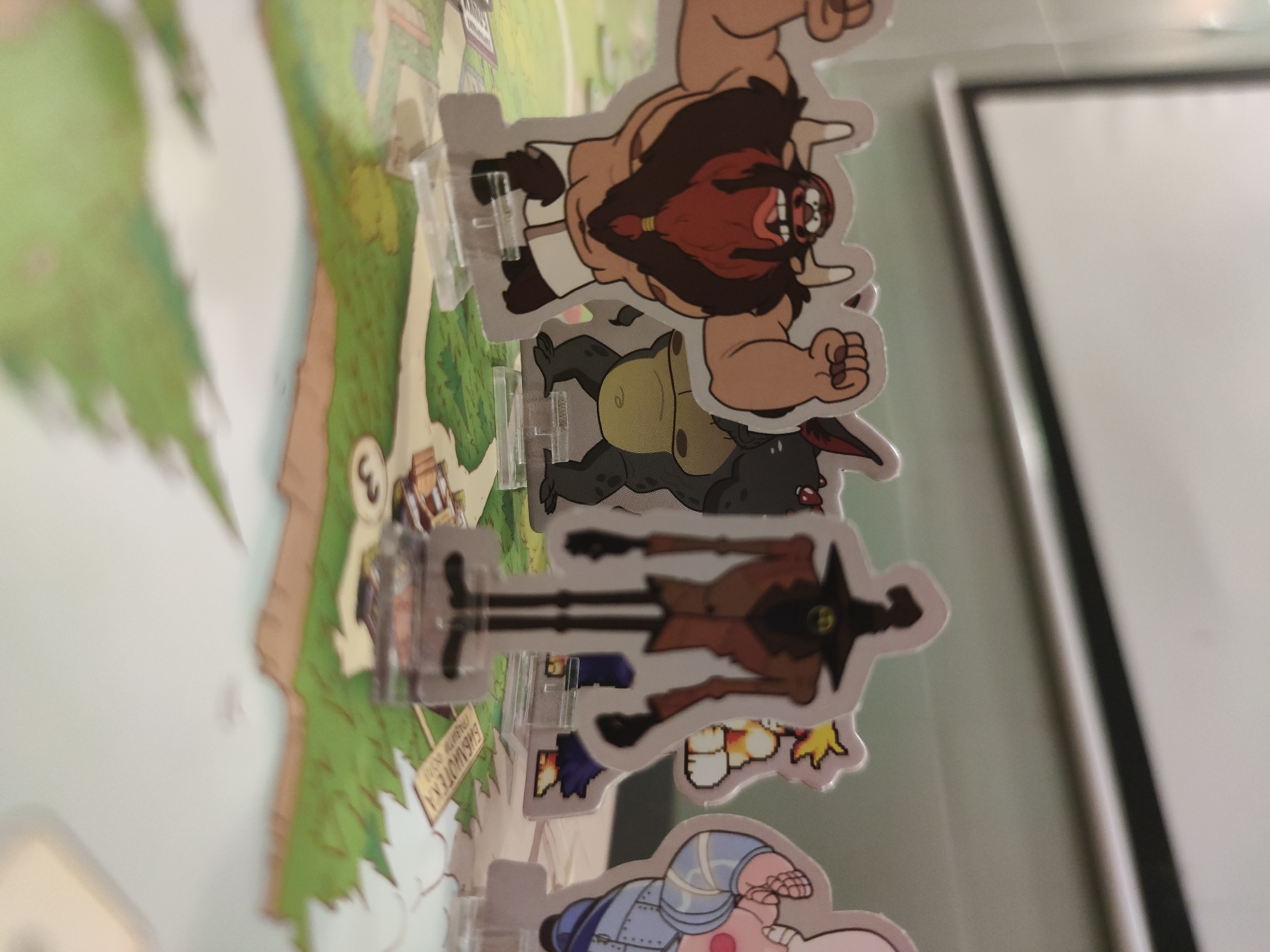 The image shows several cartoon character standees placed on a game board. The characters include a large, muscular figure with a red beard, a black and green creature, a scarecrow-like figure, and a character in blue armor. They stand on transparent plastic bases attached to a game board depicting a map with green areas and buildings. A sign labeled "Библиотека" is also visible. The photo is taken at an angle, with a white wall and a black frame visible in the background.