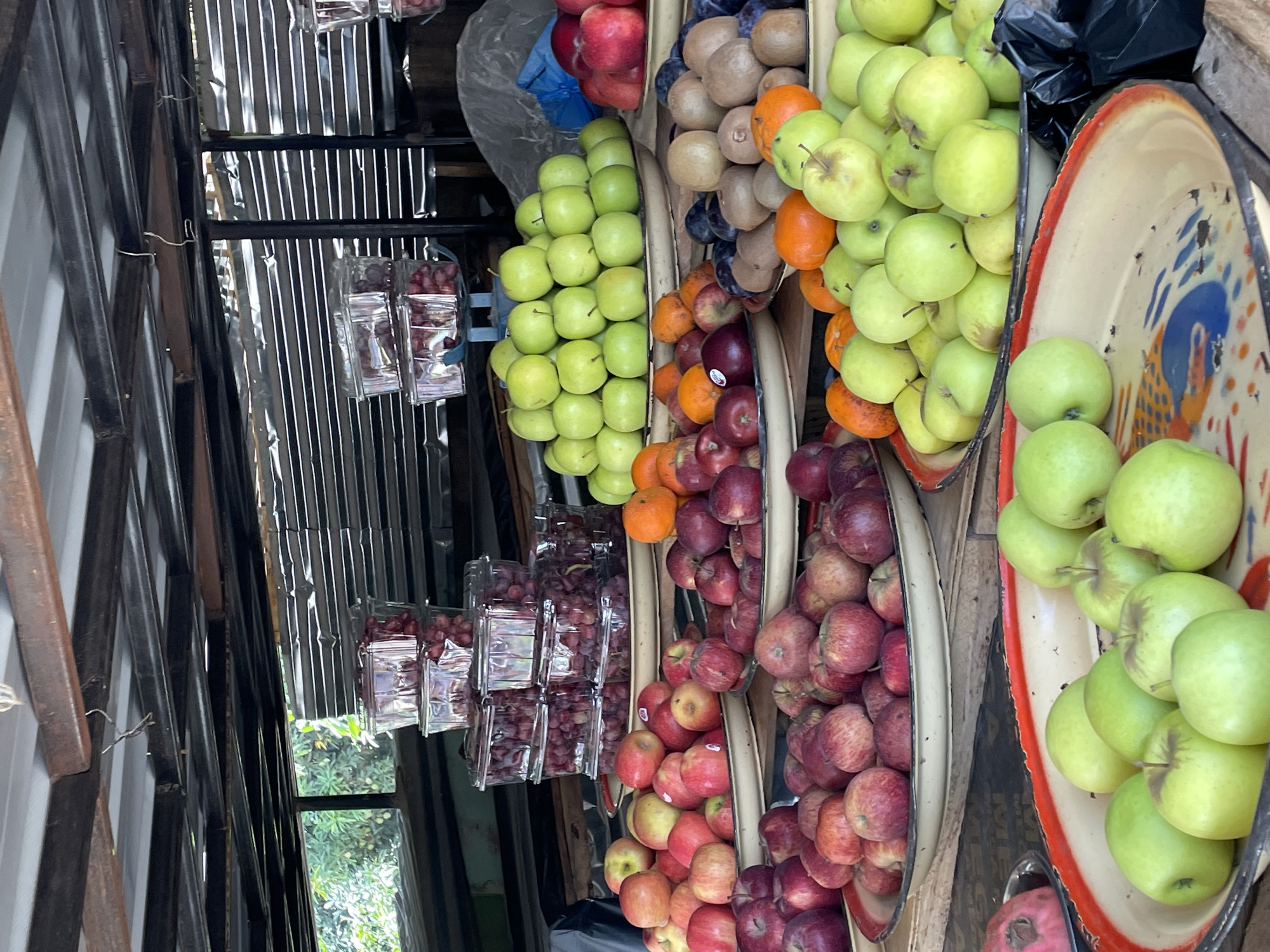The image depicts a fruit stand, likely in Jos, Nigeria, showcasing various fresh produce. Vertically displayed, from left to right, are packaged grapes, green apples, a mix of kiwis and oranges, and red apples. These are arranged in baskets and trays. The background shows a partially enclosed structure with a metal sheet wall and wooden supports. Outside the enclosure, greenery is visible through a window. The image gives a sense of a bustling marketplace.