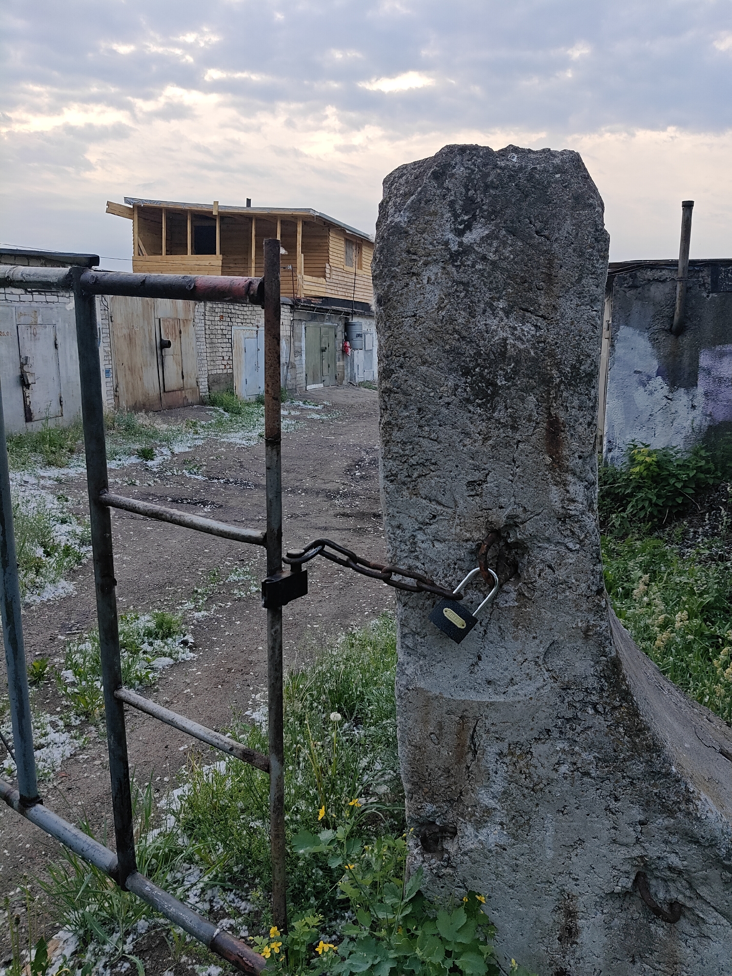 The image captures an outdoor scene, likely an alleyway or a yard behind a row of garages, set in Chaikovsky, Russia. In the immediate foreground, a rusty metal gate, appearing partially open, is secured to a large, rough concrete post. A heavy metal chain wraps around a bar on the gate and connects to a metal loop embedded in the concrete, fastened by a black padlock. The word "BULAT" is clearly visible on the padlock.
The ground beneath is a mix of dirt and gravel, interspersed with patches of green grass and weeds, and is notably covered with numerous small white specks, suggesting either fallen hail or abundant pollen.
In the midground, a long row of single-story garages extends into the distance. These structures exhibit varying materials and states of wear; some have weathered metal doors, while others are constructed from brick or have stucco finishes. Faint white text "26.01" is visible on the far-left garage door, and the letters "EPNA" are discernible on a white utility box or sign attached to a garage door further to the right.
Dominating the background is a wooden structure, possibly a makeshift dwelling or workshop, built directly atop some of the garages. This building, made of light-colored wood, features an open balcony or porch area. The sky above is overcast with scattered grey clouds, casting a soft, diffused light over the entire scene, indicative of either early morning or late afternoon. No people are present. The overall impression is one of a utilitarian and somewhat raw urban or suburban environment.