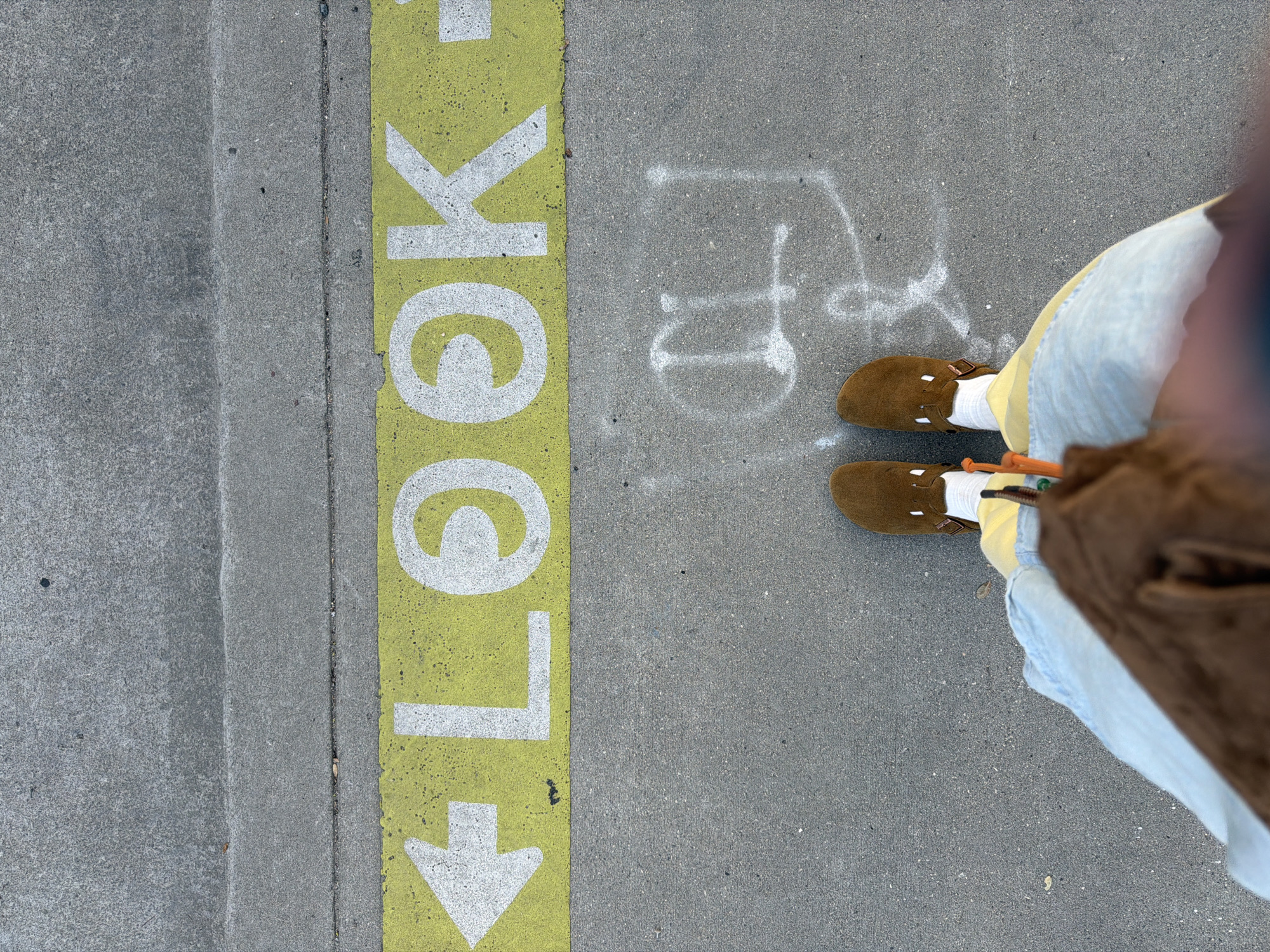 An eye-level shot looking down at a person standing on a sidewalk in West Hollywood, United States of America. The person is wearing brown suede shoes with socks, light blue pants, and a brown jacket. To the left is a yellow stripe with the word "LOOK" painted in white, with an arrow pointing downward. To the right of the yellow stripe, on the sidewalk, are faint white markings resembling the letters "FD." The sidewalk is concrete.