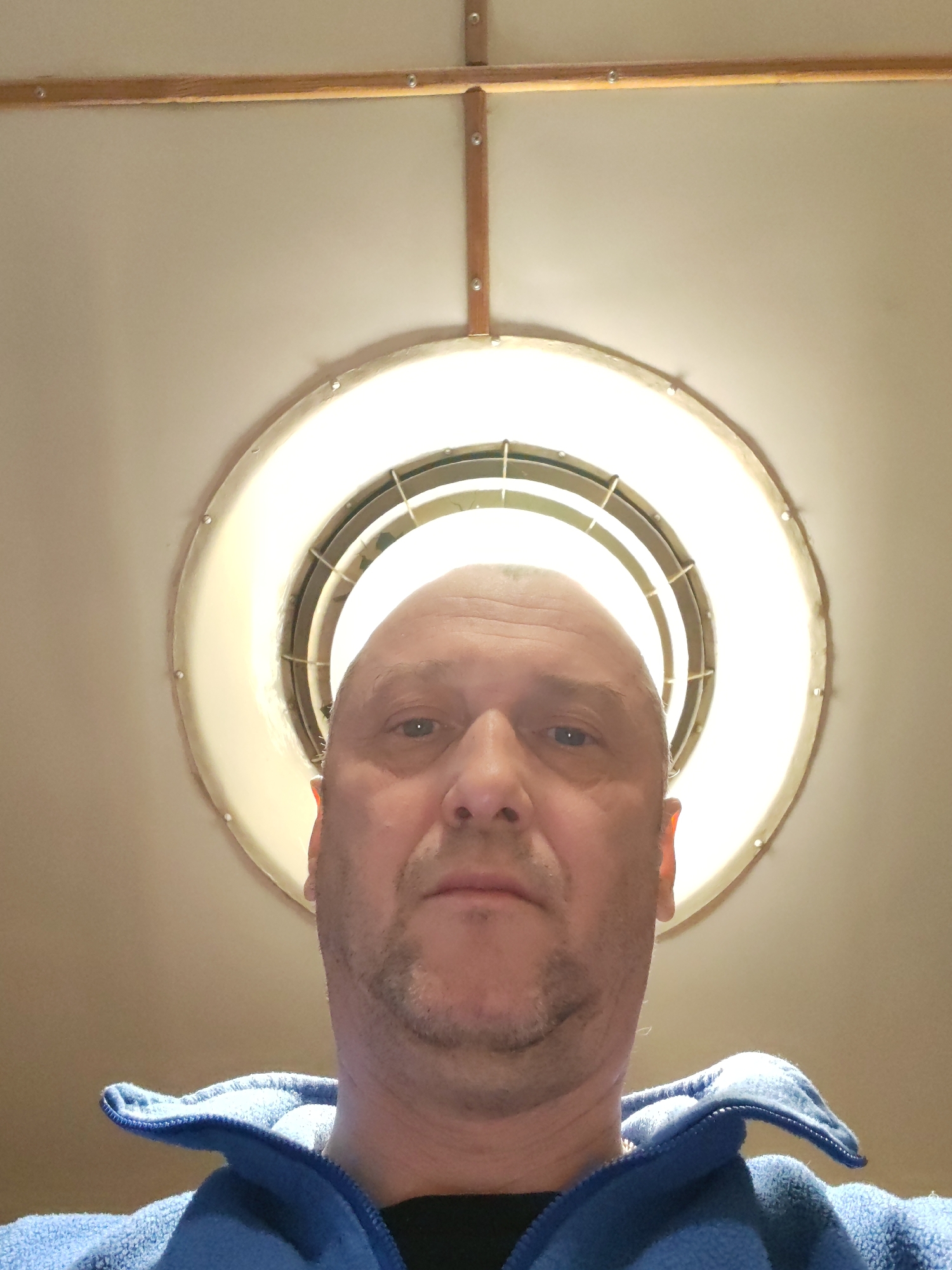 A low-angle portrait captures a bald, middle-aged man looking directly into the camera. He has light stubble on his chin and jawline, and his expression is neutral and calm. He is wearing a blue fleece jacket with a visible collar over a dark shirt.
Behind and above the man's head is a bright, circular ceiling light fixture, which appears to be a recessed or flush-mount lamp. It features a light-colored, possibly off-white or cream-colored, outer ring with visible fasteners, and internal grates or baffles. The light emanates brightly from its center. The ceiling, also a light tone, is adorned with narrow wooden strips or beams, secured by small fasteners. One wooden strip runs horizontally across the top of the frame, intersecting with another vertical strip, suggesting part of a ceiling paneling or structural design.
The indoor setting with artificial lighting does not indicate a specific time of day. The activity is simply the man posing for a direct, eye-level photograph from a low perspective. No visible text is present in the image. This scene is located in Bol'shie Mkhi, Russia.
