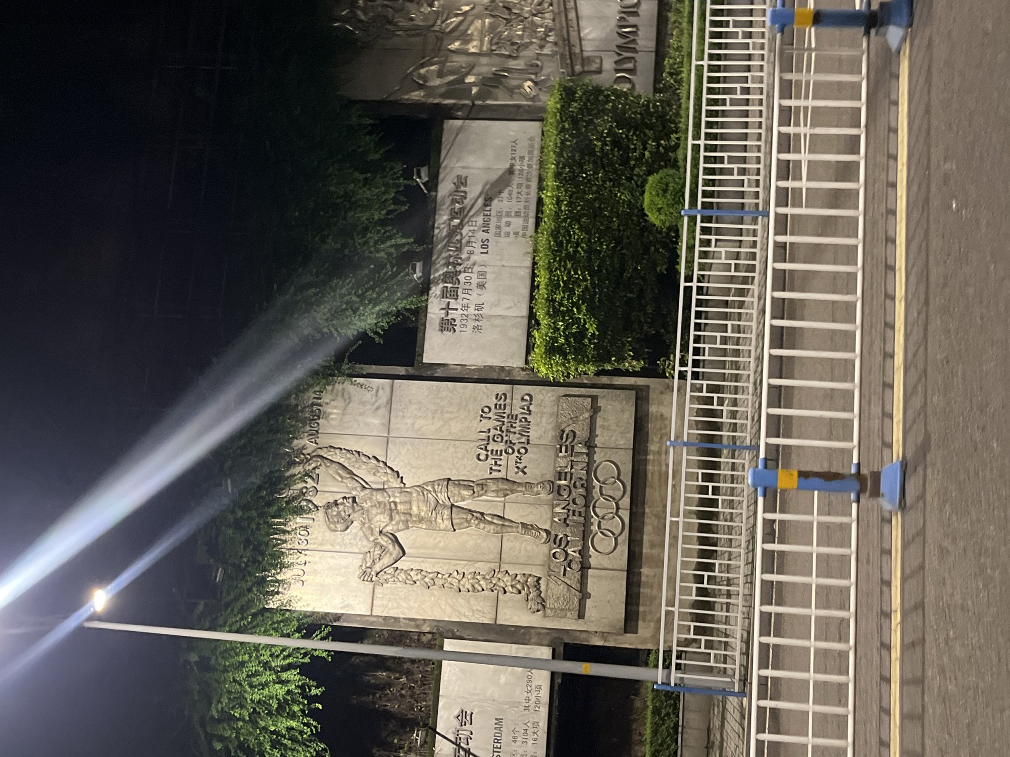The image shows a monument dedicated to the X Olympiad held in Los Angeles, California. The monument features a bas-relief of a male athlete holding a laurel wreath, with the Olympic rings at the base. Text on the monument indicates that the games took place from July 30 to August 14, 1932. A smaller plaque to the left of the main monument, also in Chinese, mentions "Amsterdam" and other details related to the Olympics. The monument is located outdoors, in what appears to be a public space, with lush green foliage overhead. In the foreground, there is a white metal fence with blue and yellow posts, and a paved road with a yellow line. The scene is illuminated by a street lamp, creating light streaks across the image. The location of Jiulongpo District, China, is associated with this monument, suggesting it is a historical marker or exhibit within the district. There are no people visible in the image.