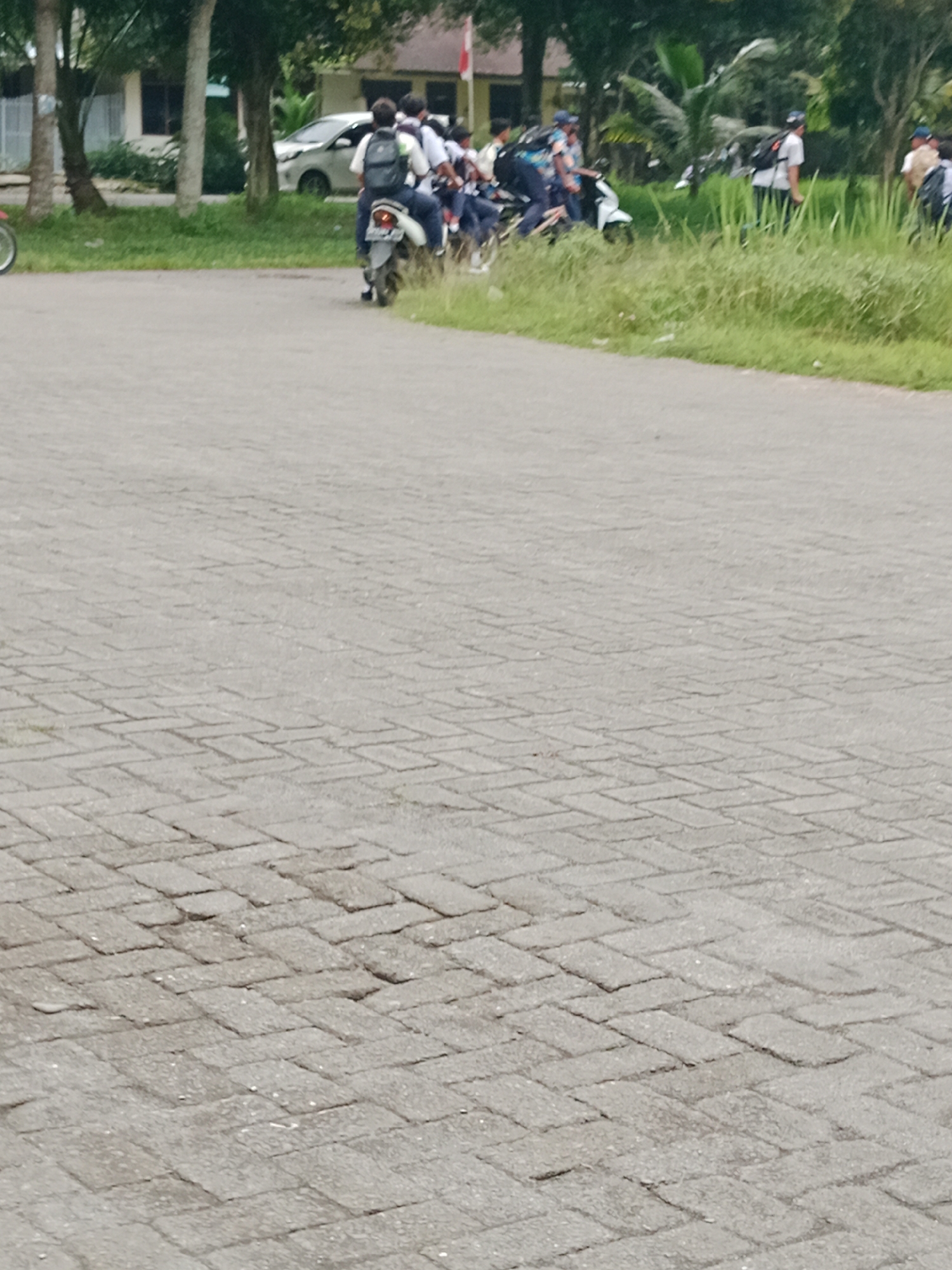 In the City of Binjai, Indonesia, a group of young people are gathered outdoors. Several individuals are seated on motorcycles, appearing to be either waiting or preparing to depart. One person in the foreground is on a white motorcycle, wearing a dark backpack and a light-colored shirt. Behind them, a line of other motorcycles is visible, with passengers also sporting backpacks. The setting appears to be a paved area, possibly a road or parking lot, bordered by lush green grass and trees. In the background, a building with a light-colored facade and a red and white flag is situated amongst the trees. Another white car is parked to the left of the building. The sky is not visible, and the overall lighting suggests it is daytime.
