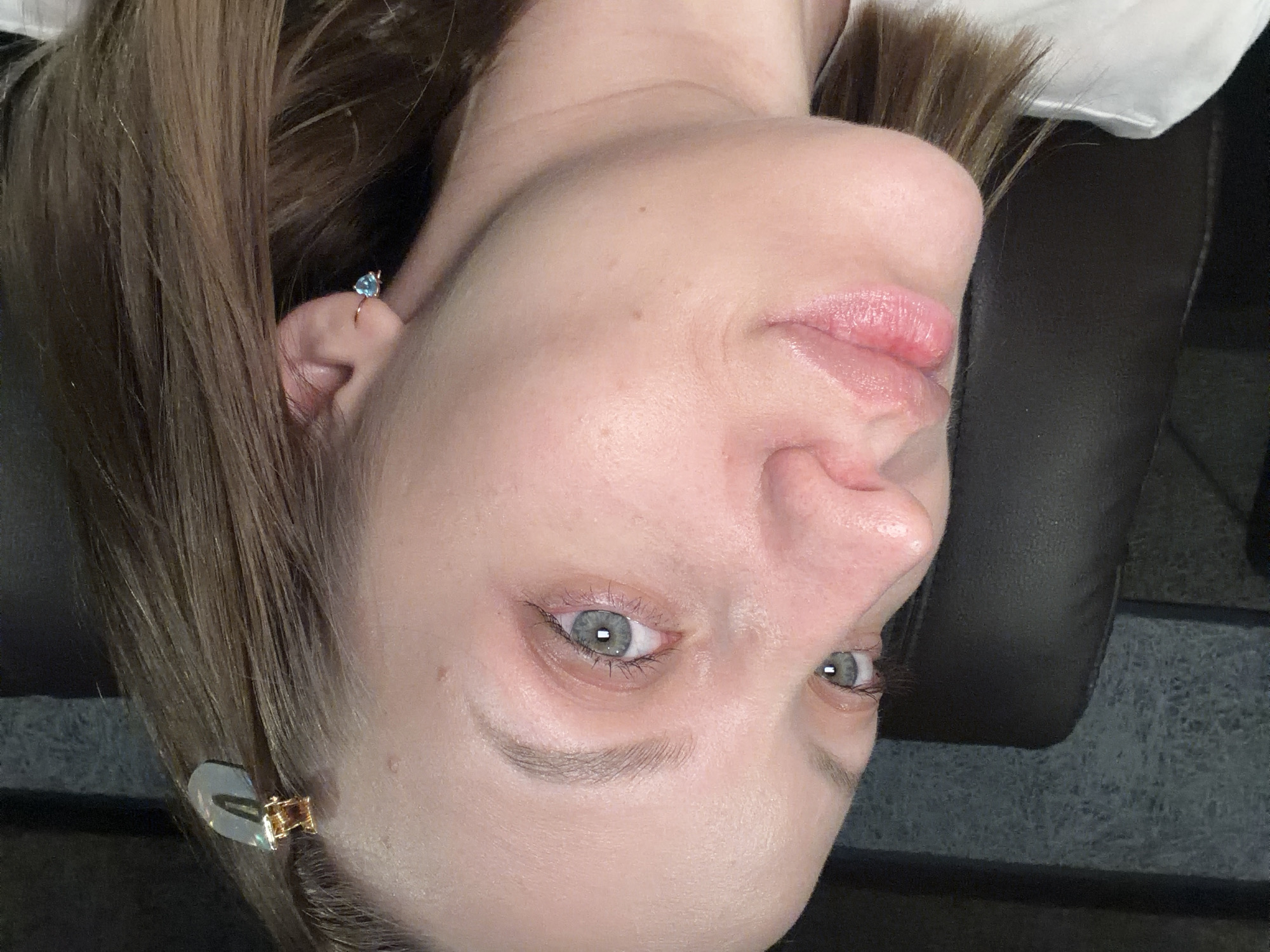 A close-up, low-angle, indoor photograph shows a young Caucasian woman's face and upper torso. She is facing left, with her head slightly tilted down. The woman has fair skin, light brown hair styled with a golden hair clip, and light blue eyes. She is wearing a light blue heart-shaped earring. Her lips are slightly parted and appear glossy. A dark leather chair and a textured wall are visible in the background. The image was taken in Klin, Russia.