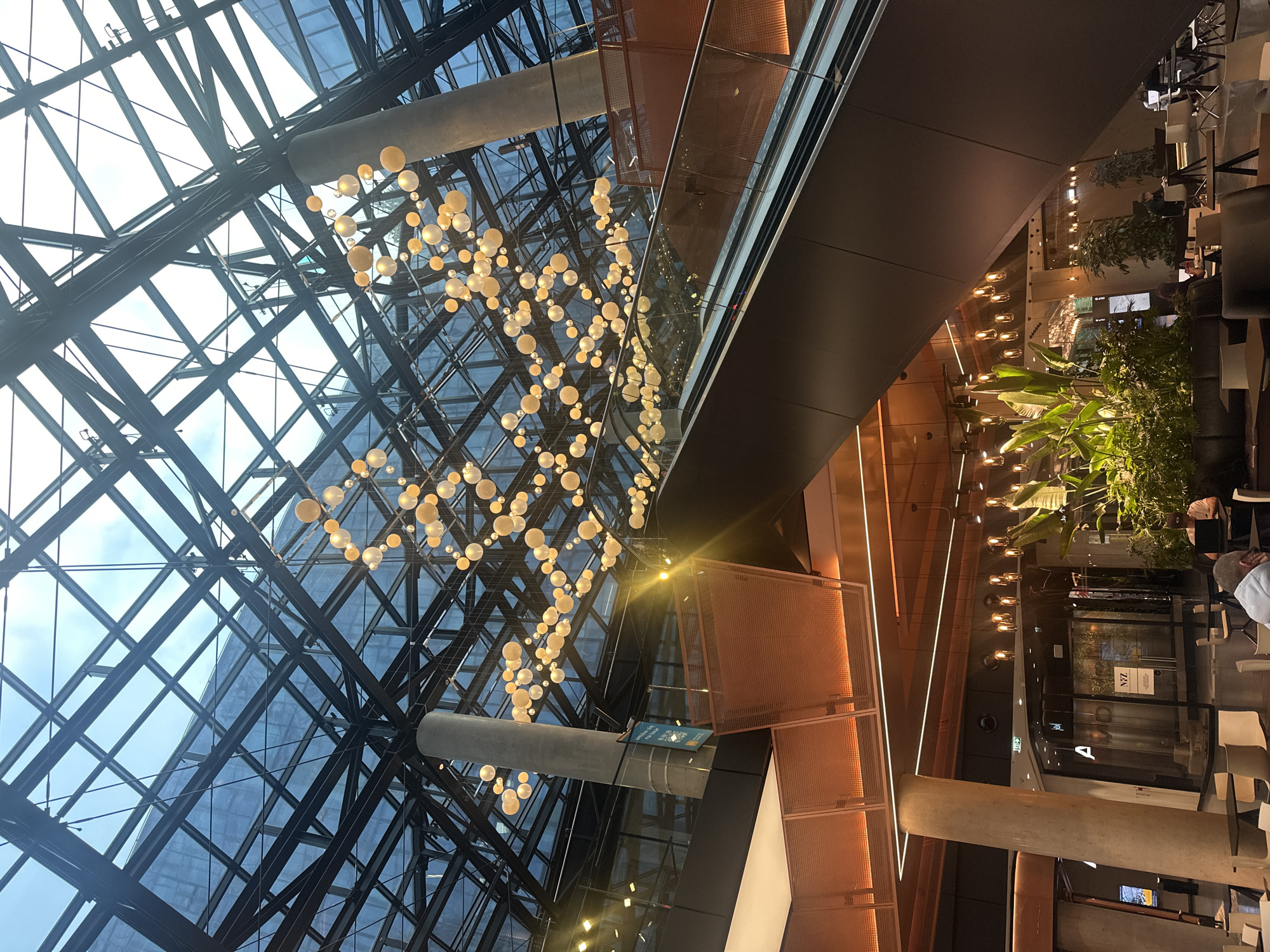 The image shows the interior of a modern building with a large, glass, geometric ceiling structure. Hanging from the ceiling are numerous spherical, yellowish lights, clustered together. An escalator is visible on the right side of the frame, its dark metallic structure contrasting with the lighter elements of the interior. In the lower section of the image, there are tables and chairs, suggesting a seating or dining area. A large plant is situated to the left of the escalator, adding a natural element to the man-made environment. The overall impression is of a spacious, contemporary atrium. The scene is located in Warsaw, Poland.