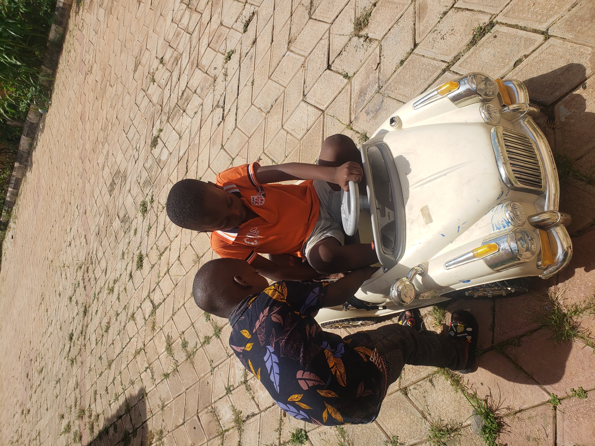 Two young Black boys are playing with a white toy car on a paved courtyard in Sabon Tasha, Nigeria. The boy on the left, wearing a black shirt with a colorful leaf print and dark pants, is leaning over the toy car, appearing to be pushing or adjusting it. The boy on the right, dressed in an orange polo shirt and grey shorts, is seated in the toy car, his hands on the steering wheel. The ground is paved with light brown interlocking bricks, with patches of grass growing in the gaps. Lush green vegetation is visible in the background along the edge of the courtyard. The scene is illuminated by bright daylight, casting shadows on the ground.