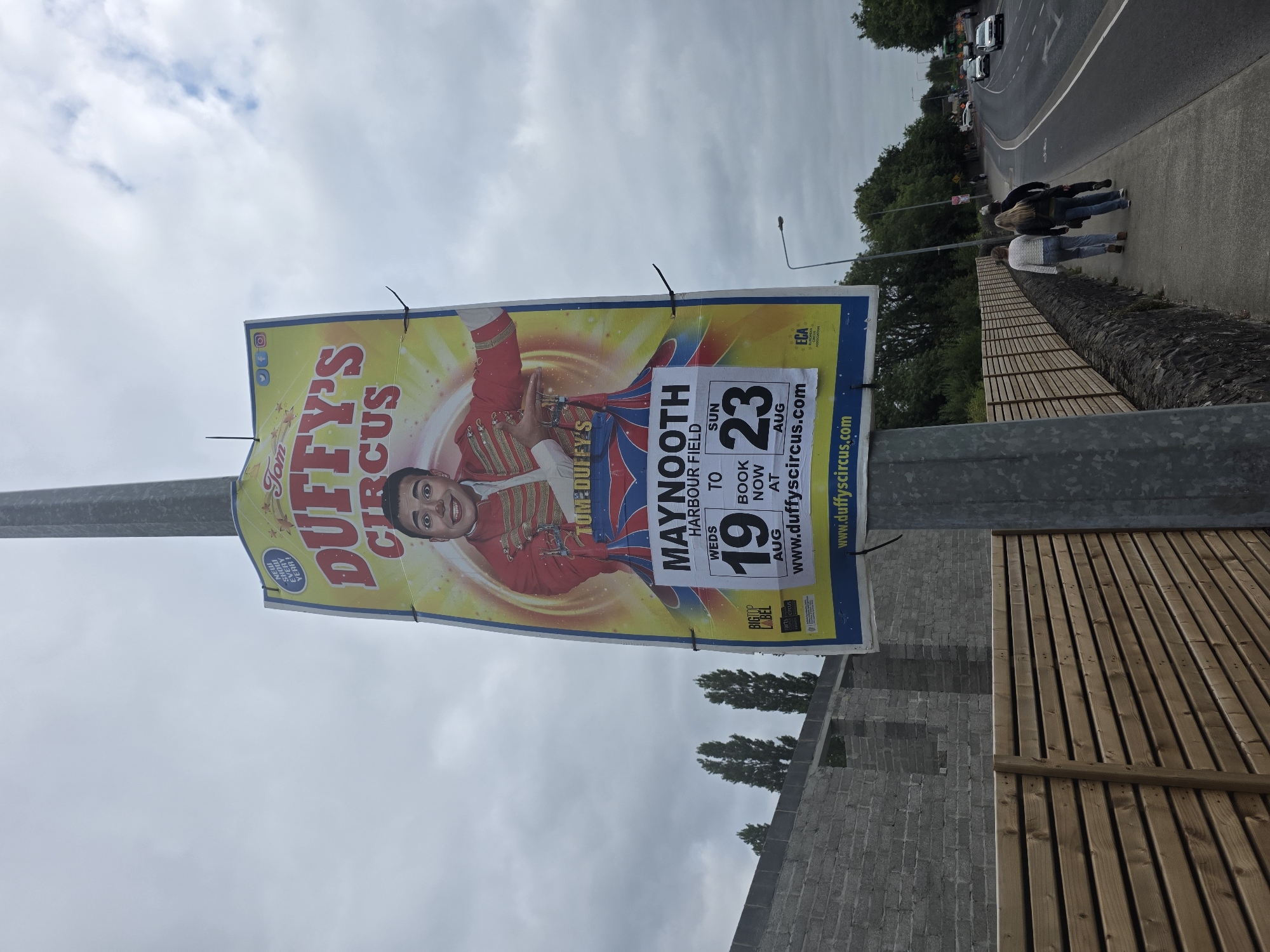 A tall, vertical circus poster is affixed to a metal pole on a cloudy day. The poster advertises "Tom Duffy's Circus" with a central image of a smiling man in a red circus costume. Below the image, it states "MAYNOOTH HARBOUR FIELD" and provides dates "WED 19 AUG to SUN 23 AUG" for performances, with a call to action to "BOOK NOW AT WWW.DUFFYSCIRCUS.COM". The poster also features smaller logos such as "BIGTOP LABEL" and "ECA".
To the left of the pole, a grey brick wall is visible, partially under construction. In the foreground, a wooden slatted fence runs along a stone wall. A paved footpath is to the right of the fence, with three people walking away from the camera. Beyond the footpath, a road curves to the right, with several cars driving on it. Green trees and foliage line the side of the road. The overall setting appears to be an outdoor area in or near the city of Maynooth, Ireland, indicated by the poster's text.