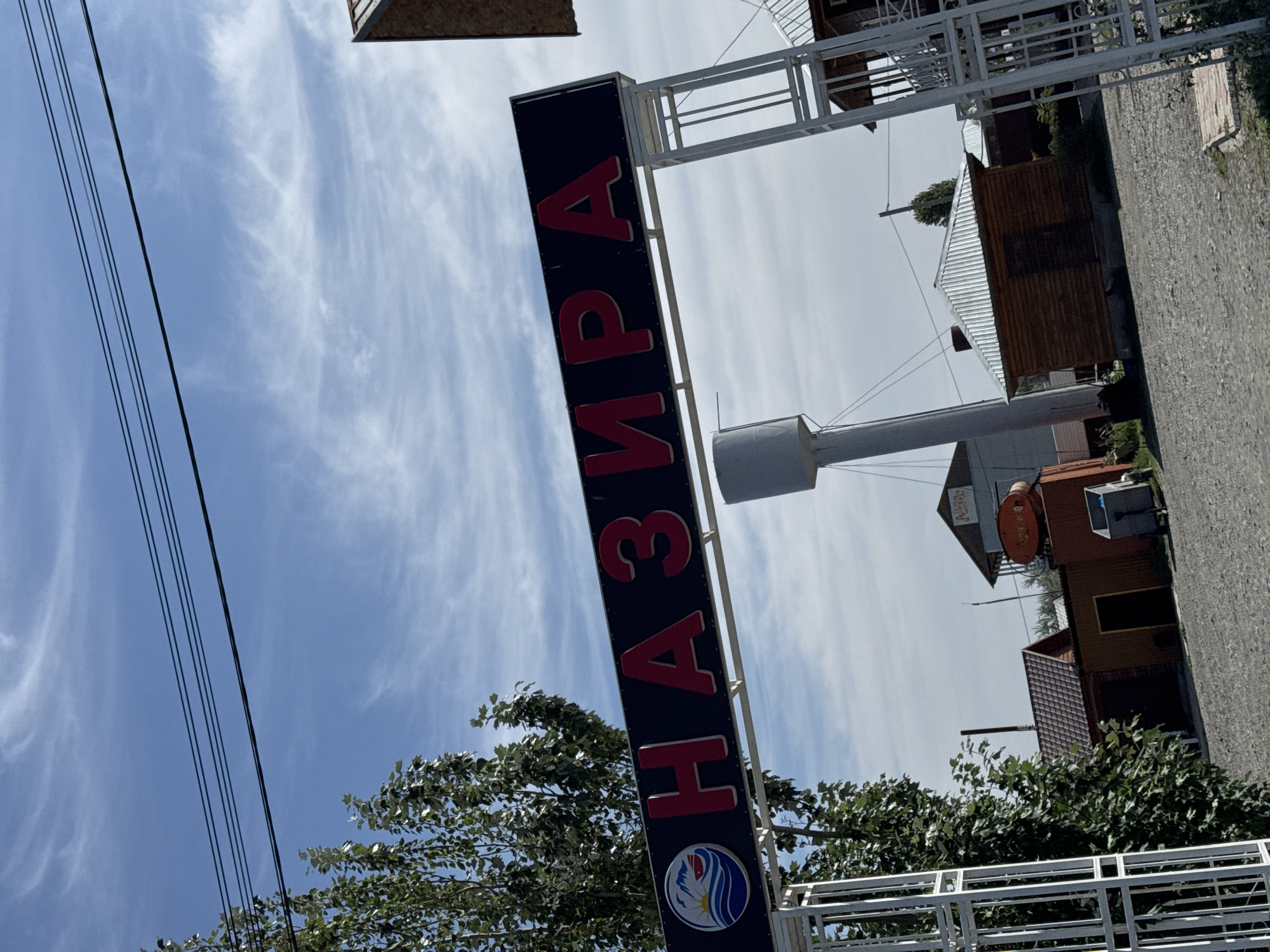 A wide-angle, low-angle outdoor shot features a sign with red Cyrillic lettering that reads "НАЗИРА" on a dark blue background, mounted on a white archway. To the left of the sign is a circular logo with a stylized wave and mountain landscape. Behind the archway, a tall, slender white water tower is visible. Several small, rustic wooden buildings with metal roofs are scattered across a gravel-covered area. Trees with green foliage are present on the left side of the frame. In the background, telephone wires are strung across a bright blue sky with wispy white clouds. The overall scene suggests a rural or semi-rural location, potentially a resort or a small settlement. The text on the sign is likely the name of the place.