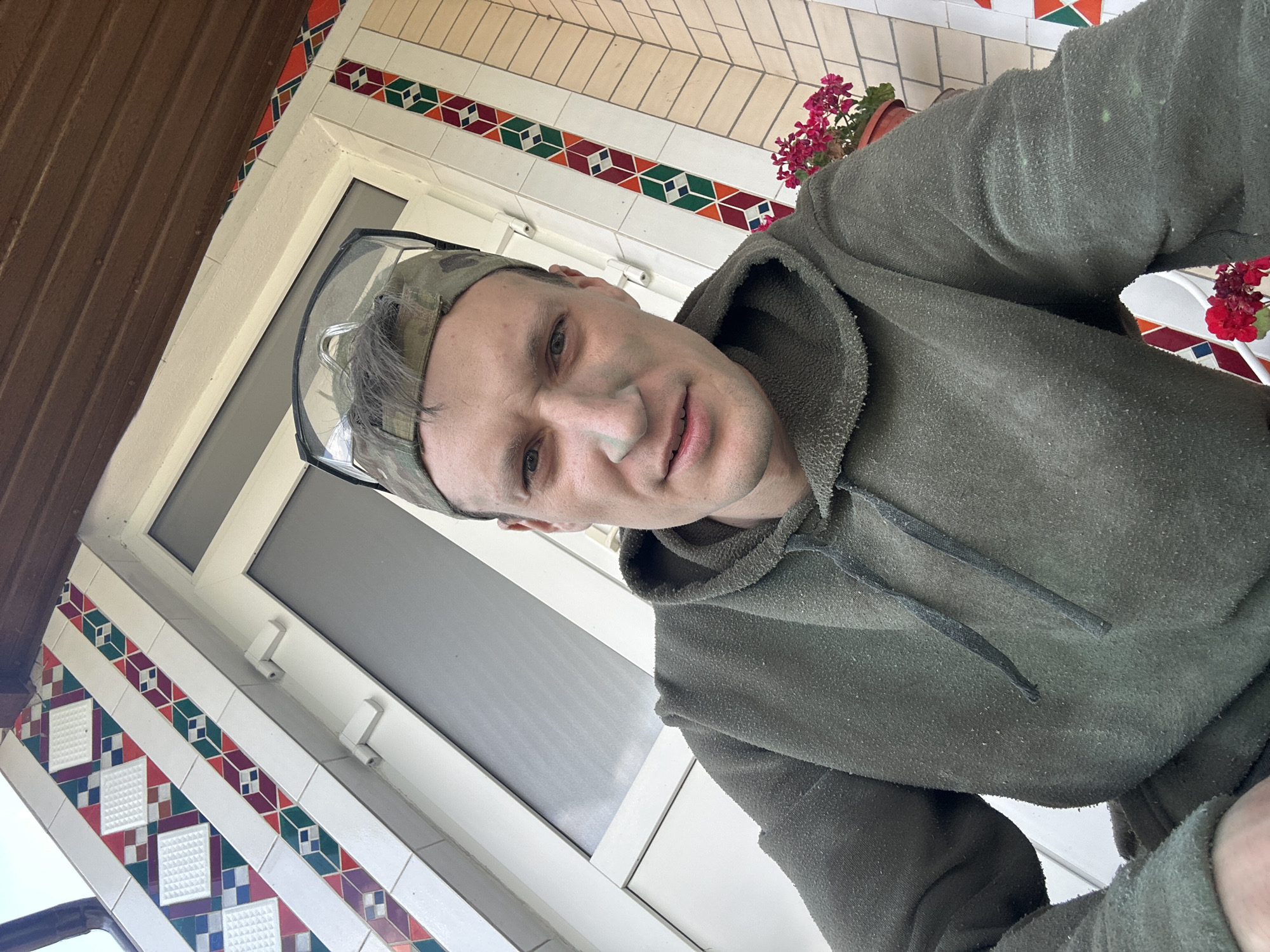 A man wearing a green hoodie and a camouflage baseball cap with safety goggles on top of it is pictured from the chest up. He has green dust or paint on his face, particularly around his nose and cheeks. The man is looking directly at the camera with a slight frown. In the background, there is a white building with decorative tiles featuring red, green, and purple geometric patterns. There are also some potted flowers with red blossoms visible. The image appears to be taken outdoors, possibly on a porch or near the entrance of a building. The lighting suggests it is daytime. The general setting appears to be residential.