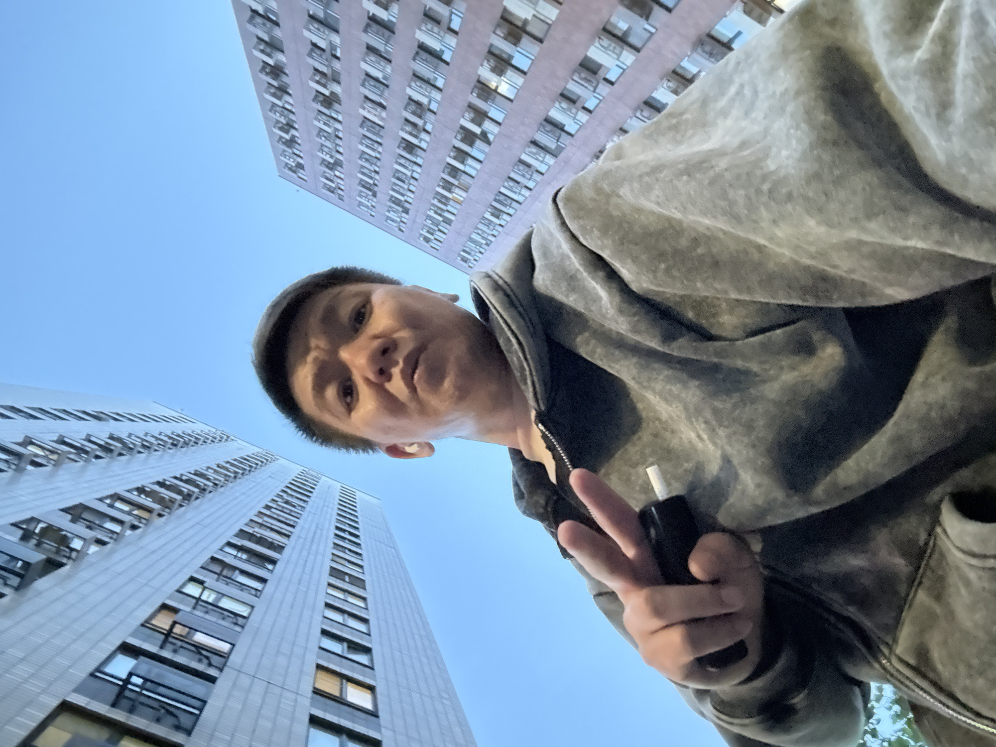 A man wearing a gray hoodie and holding a black electronic cigarette in his right hand poses for a low-angle selfie. He makes a "peace" or "victory" sign with his left hand. Two tall buildings are visible in the background, stretching towards the blue sky. The architecture suggests the location is Moscow, Russia.