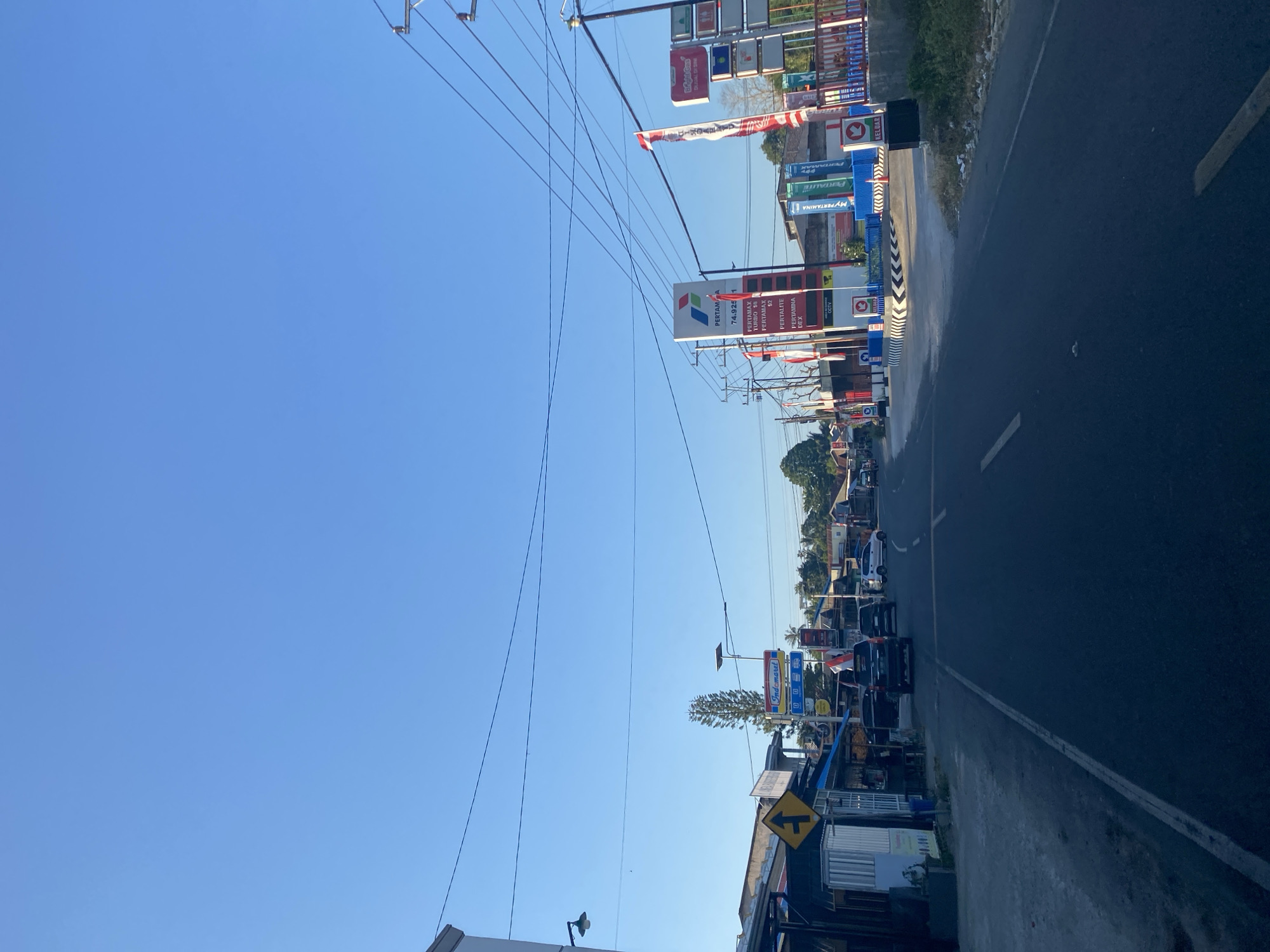 This is a photograph taken from a street in Palambarae, Indonesia, looking towards a Pertamina gas station. The sky is clear and blue. On the left side of the road, there are several small shops with awnings, and a yellow directional sign. Further down the road, the Pertamina gas station is prominent, with its red and white logo and price boards displaying fuel types and costs, including Pertamax, Pertalite, and Pertamina Dex. Several cars are visible on the road and parked at the gas station. A few people are standing near the shops. The environment appears to be a semi-urban area with a mix of commercial establishments and residential buildings in the background. Electrical wires are visible strung across poles.