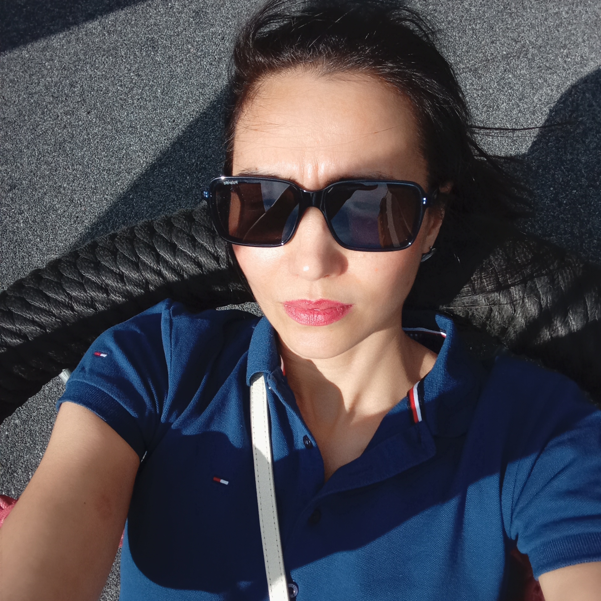 A woman wearing sunglasses and a blue polo shirt with a white strap is lying down. The background is a textured gray surface with a dark rope-like element. The location is unknown and cannot be determined from the image. The city of Pervomaysky, Russia is not mentioned or discernible.