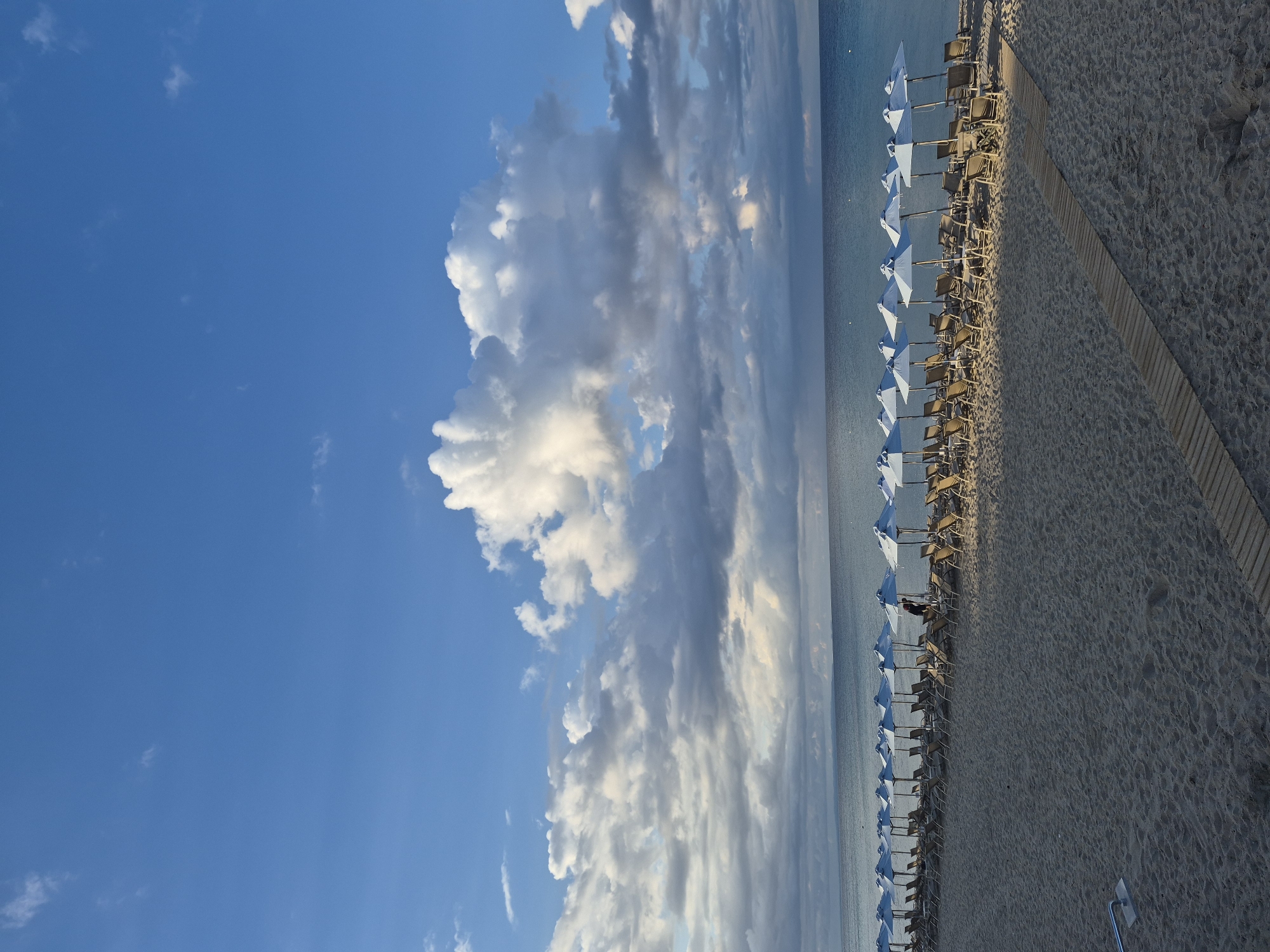 A wide, empty beach in Nea Kallikratia, Greece, is pictured under a bright blue sky with dramatic, fluffy white clouds. The sand is light-colored and textured, with a wooden walkway leading diagonally from the foreground towards the sea. A long, orderly row of beach chairs and white umbrellas are set up along the shoreline, mostly unoccupied, with a single person visible between two umbrellas in the middle of the frame, appearing to be adjusting one of them. The calm, turquoise sea stretches to the horizon, meeting a hazy blue sky. The overall impression is one of serene tranquility and readiness for a day at the beach.
