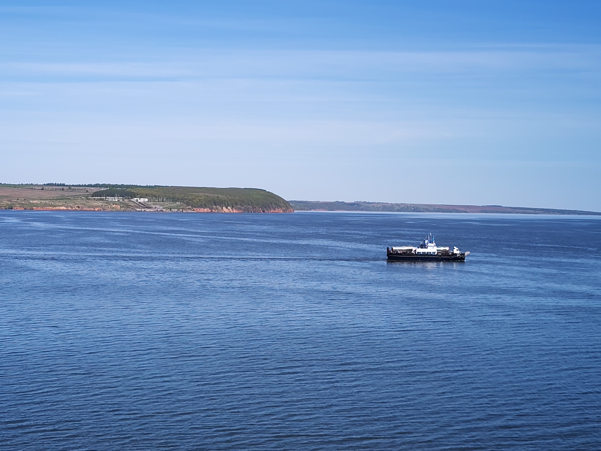 The scene depicts a body of water, presumably a river or lake, under a clear, slightly clouded sky. A ferry navigates the water, leaving a visible wake. In the background, a landmass with a distinctive reddish-brown cliff face topped with green vegetation is visible. The location is Икское Устье, Russia.