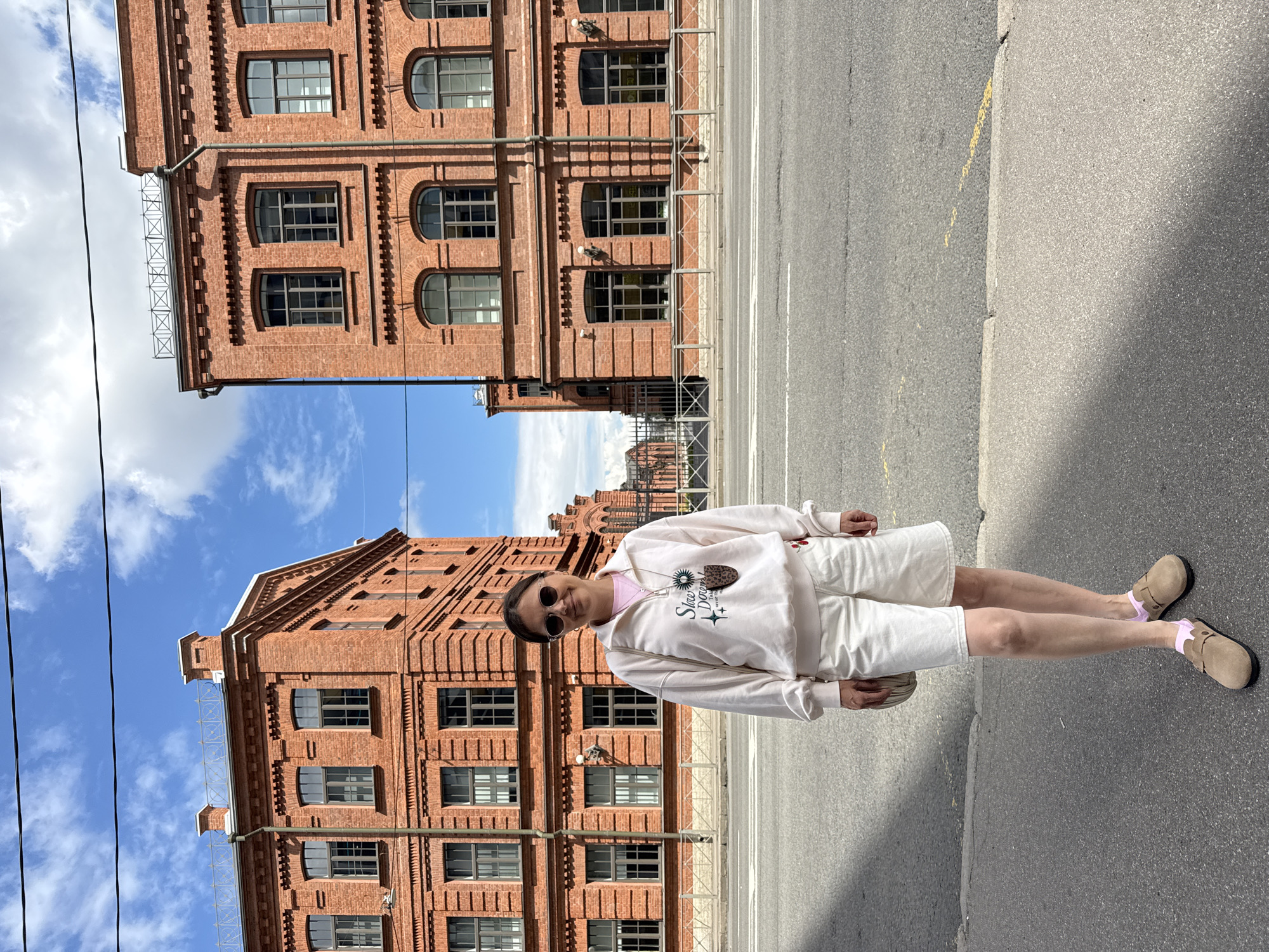 A woman stands on a sidewalk in front of two imposing brick buildings in Saint Petersburg, Russia. The buildings are typical of 19th-century industrial architecture, with numerous multi-paned windows and decorative brickwork. The woman is wearing a light-colored sweatshirt, white shorts, and tan clogs with pink socks. She has sunglasses on and a necklace with a leopard-print pendant. The sky is blue with scattered white clouds. The foreground shows a paved street with lane markings and a sidewalk. The overall impression is one of an urban setting on a clear day.