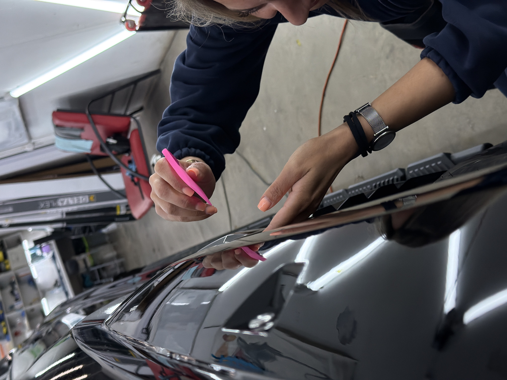 A person is shown applying a vinyl wrap to a black car. The person is wearing a navy blue long-sleeved shirt and a watch. The person is using a pink squeegee. The car appears to be in a garage, and there are tools and equipment visible in the background. There is no information available to confirm the location of this garage as Krasnoyarsk, Russia.