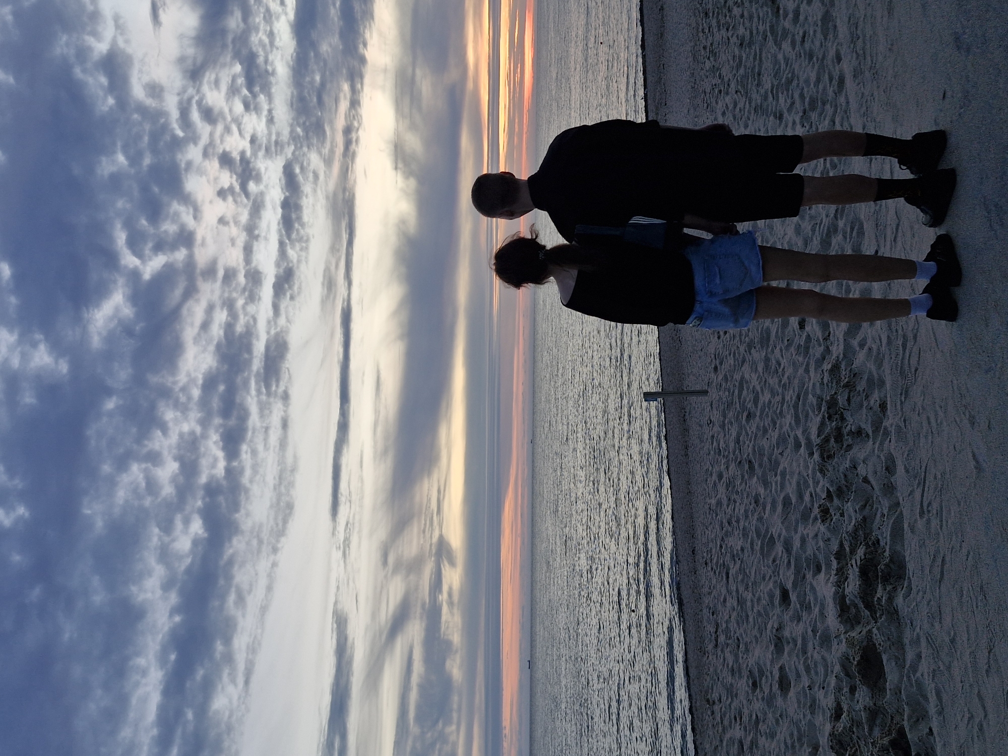 Two individuals, a female and a male, stand facing away from the camera, looking out at the sea and sky from a sandy beach. The female is on the left, wearing a black off-the-shoulder top and denim shorts. Her hair is pulled back into a ponytail. The male is on the right, wearing a black t-shirt and black shorts, with dark socks and shoes. The sky is filled with dramatic clouds, ranging in color from deep blue to light grey and white, with streaks of orange and pink visible near the horizon, indicating sunset or sunrise. The sea is calm, with a few distant ships visible on the horizon. The sand on the beach is textured with footprints. A thin wooden post is visible in the sand to the right of the couple. This scene is likely the Curonian Spit, a narrow, sandy peninsula that is part of Klaipėda, Lithuania, known for its beaches and natural beauty.