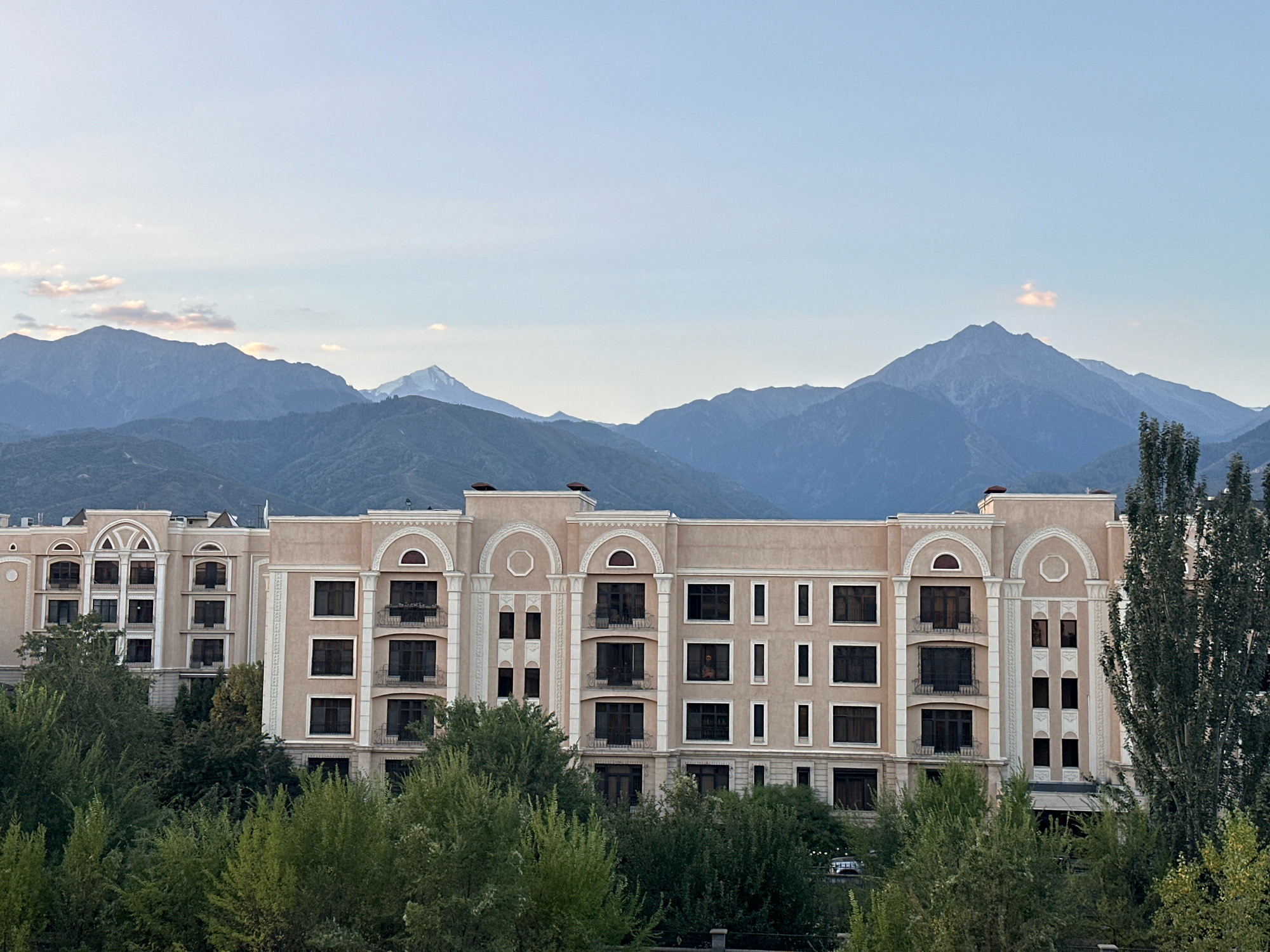 The image shows a large, light-colored apartment building with a tiered facade, arched windows, and balconies. It is surrounded by lush green trees in the foreground and middle ground. In the background, several layers of mountains are visible, with the sky above showing a gradient of light blue. One of the mountain peaks in the distance is snow-capped. The building appears to be modern in design, with intricate architectural details such as decorative molding and columns. The presence of trees and mountains suggests a location on the outskirts of a city, possibly in a region with a mountainous landscape. Based on the mountainous backdrop, this scene is likely located in Almaty, Kazakhstan, a city known for its proximity to the Tian Shan mountain range.