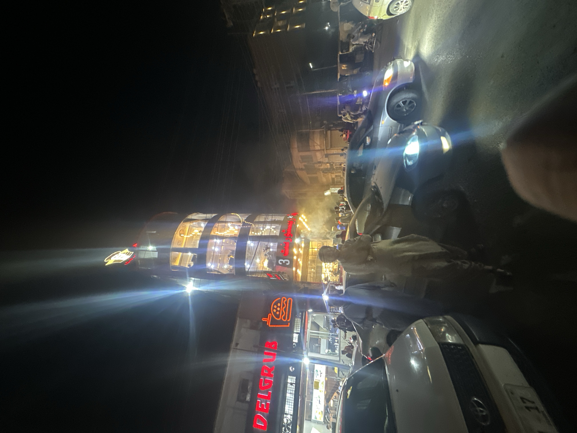 The image captures a nighttime street scene in Quetta City Tehsil, Pakistan. A multi-story cylindrical building is illuminated, with the name "DELGRUB" displayed in red neon lights on its lower section. The top of the building appears to have a sign. Cars are parked on the side of the road, and people are visible on the sidewalk. Overhead power lines run above the street. The lighting is uneven, creating bright spots and shadows.