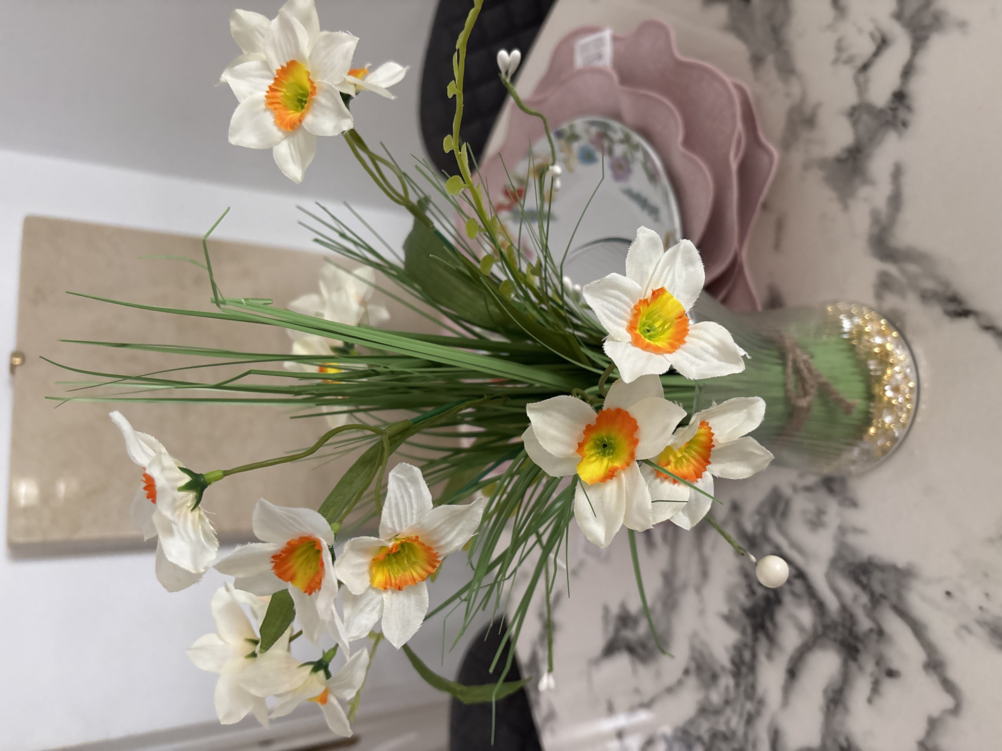 In a close-up shot, a clear glass vase holding a bouquet of artificial white daffodils with orange and yellow centers sits on a marble countertop. The vase is tied with twine and contains small gold decorative beads at the bottom. The daffodils are arranged with green artificial grass, creating a full and vibrant display. In the background, out of focus, a stack of pink ruffled placemats and a small decorative plate with a floral pattern are visible. The overall scene suggests an indoor setting, possibly a dining table or a sideboard. The image does not contain any information that would allow for identification of its location in Orihuela, Spain.