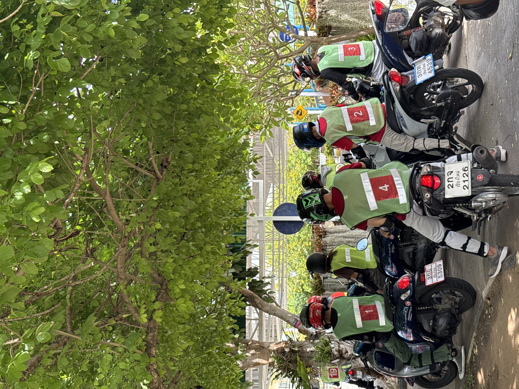 The image shows a group of people on motorbikes, likely in the city of Nong Phueng, Thailand. The individuals are wearing numbered vests (1, 2, 3, 4) and helmets. The motorbikes are parked next to each other, and license plates are visible on the vehicles. A tree is located to the left, providing some shade to the area. A building with a white-framed structure is visible in the background.
