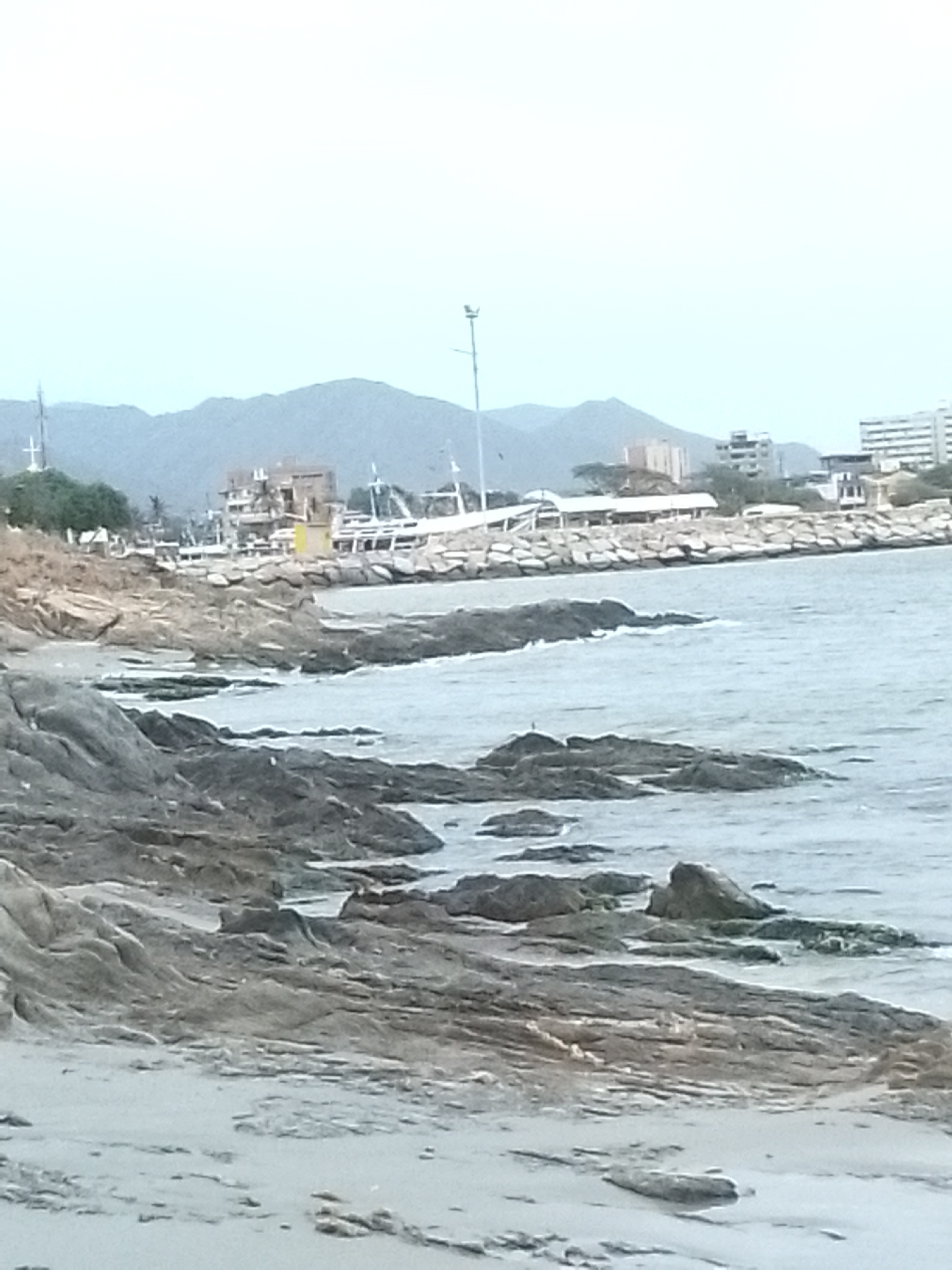 This image depicts a coastal scene in Juan Griego, Venezuela. In the foreground, there is a rocky shoreline with gray, weathered rocks extending into the sea. The water is calm, with small waves lapping against the rocks. In the background, there is a breakwater constructed with large rocks, extending out into the bay, and a small complex of buildings and parked boats. Behind the breakwater, there are buildings of varying sizes and styles, consistent with a coastal town. In the far background, there are low-lying mountains or hills under an overcast sky. There are no people immediately visible.