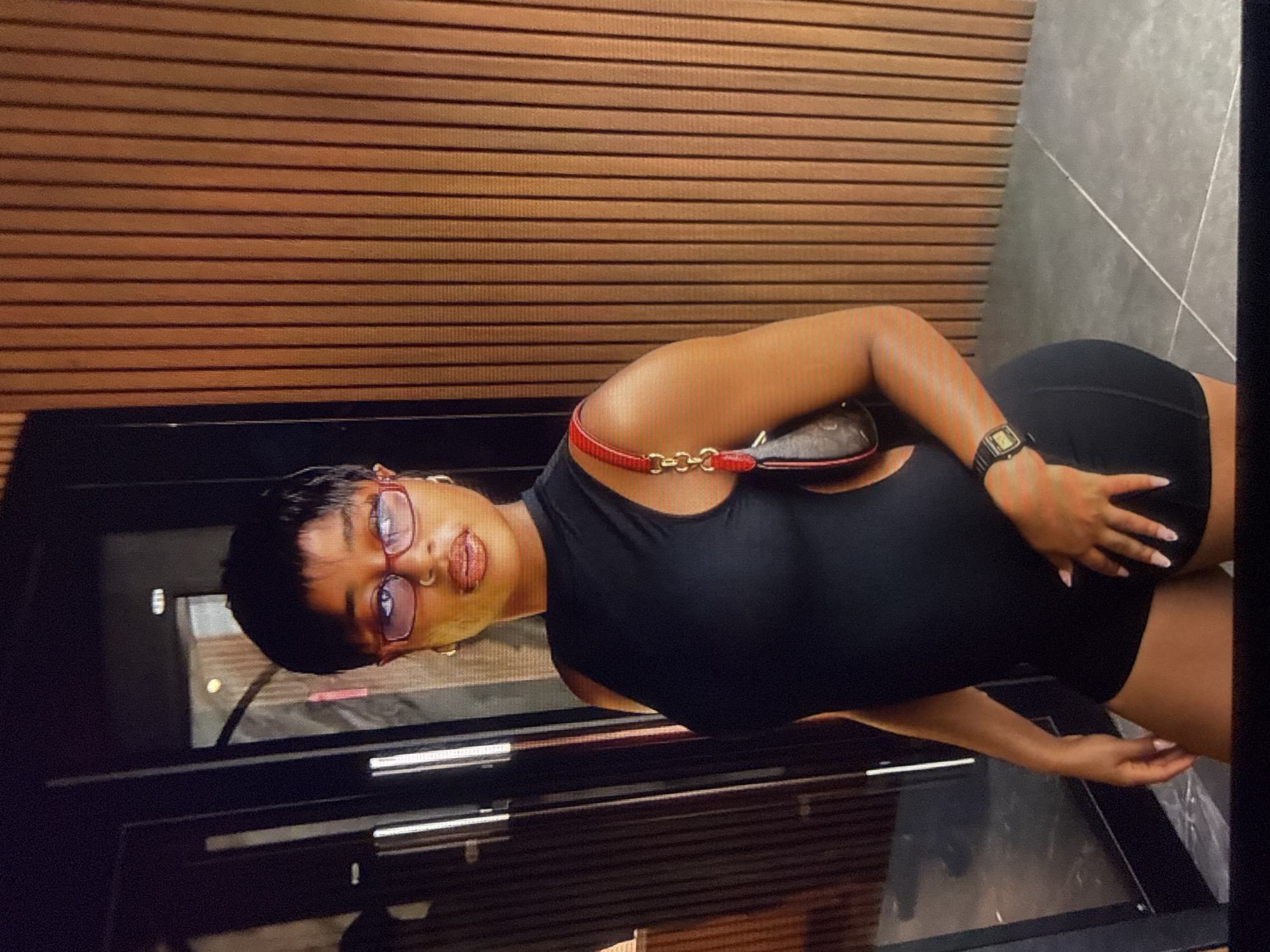 A Black woman with short black hair and glasses is standing and posing for a picture. She is wearing a black sleeveless outfit and carrying a small brown and red purse. The background is a wooden wall. The scene appears to be located in a modern indoor setting. There is no information to indicate the location is Abuja, Nigeria.
