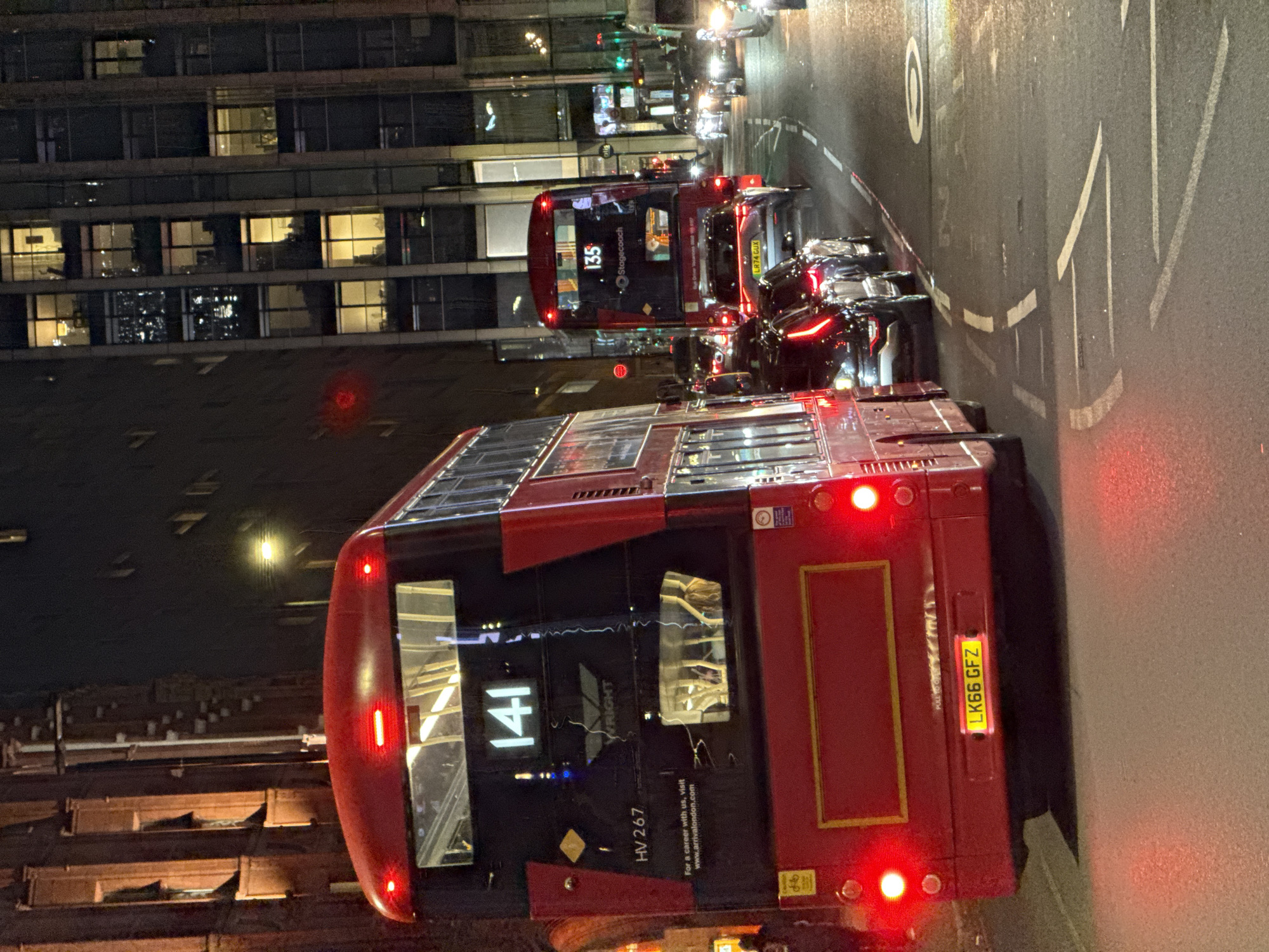 The image captures a nighttime street scene in London, United Kingdom, featuring two red double-decker buses. The foremost bus, marked with route number "141," has the license plate "LK66 GFZ." The second bus, partially visible behind it, displays the route number "135" and the operator "Stagecoach" with the license plate "LR74 GUX." Both buses are surrounded by other vehicles and pedestrians. The backdrop consists of a tall building with illuminated windows. Street markings and signs are also visible on the road.
