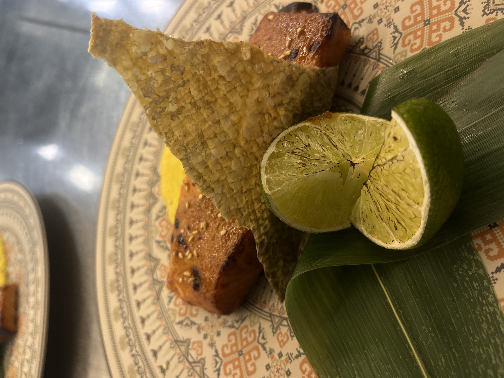 The image depicts a plate of food, potentially in a restaurant or home setting in Sofia, Bulgaria, given the request context. On a patterned plate, there are pieces of grilled fish or tofu coated in sesame seeds, a large fried fish skin, a bright yellow sauce, and a cut lime placed on a long green leaf. Another plate with the same food is slightly visible on the left side. The surface below the plates appears to be metallic or stainless steel.