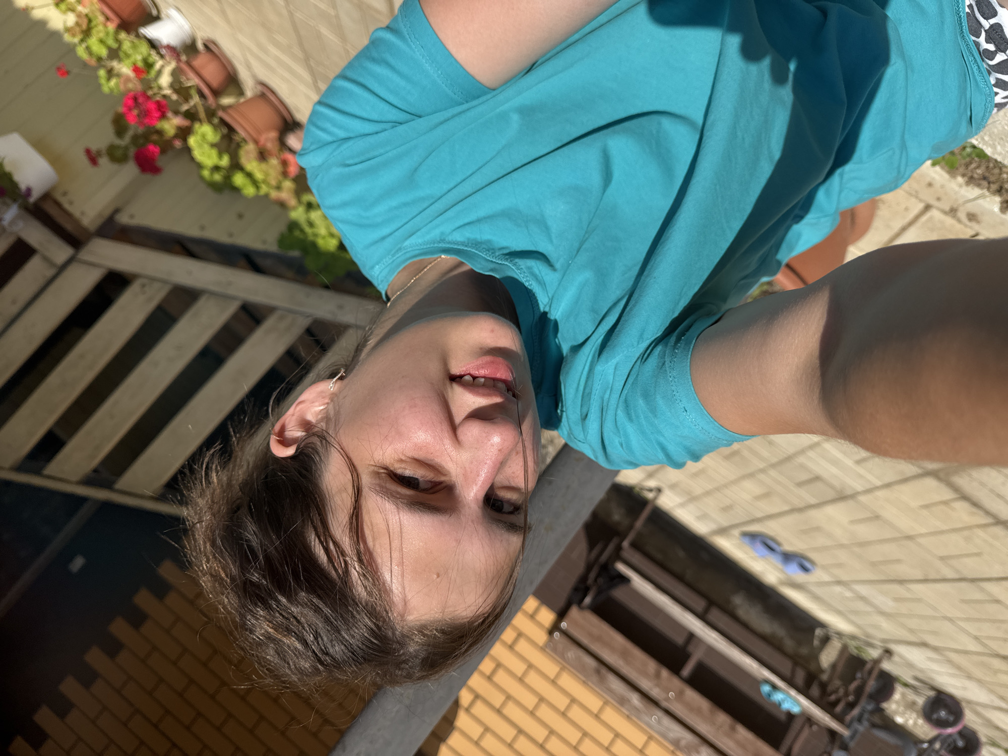 The image shows a young woman with brown hair and brown eyes, wearing a turquoise t-shirt and what appears to be patterned shorts. She is outdoors, in what seems to be a backyard or patio area, with a wooden structure and brickwork visible in the background. Potted plants with red and green foliage are also present. The lighting suggests it is daytime. The image is taken from a high angle, looking down at the woman. There are no specific details that place the scene in Vereshchagino, Russia.