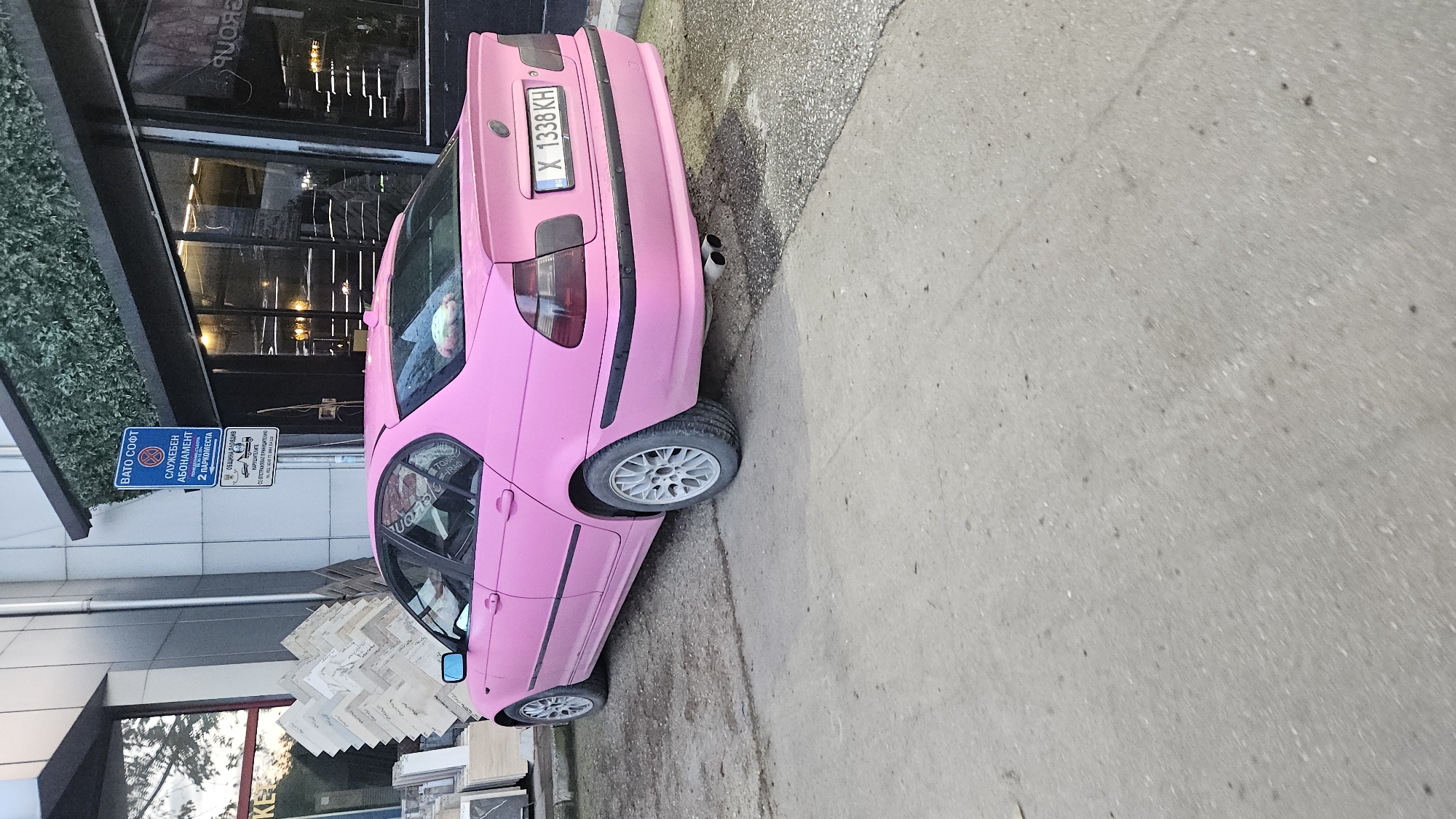 The image shows a bright pink car parked on the side of a street. The car's license plate reads "X 1338 KH." The car is next to a building with a store that has large window displays and what appears to be tile samples in front of the building. A sign on a pole indicates "BATO SOFT," "СЛУЖЕБЕН АБОНАМЕНТ 2 ПАРКОМЕСТА", and "ОБЩИНА ПЛОВДИВ НАРУШИТЕЛИТЕ СЕ ОТСТРАНЯВАТ ПРИНУДИТЕЛИО," suggesting that the location is in the city of Plovdiv, Bulgaria. The street and sidewalk are made of concrete.